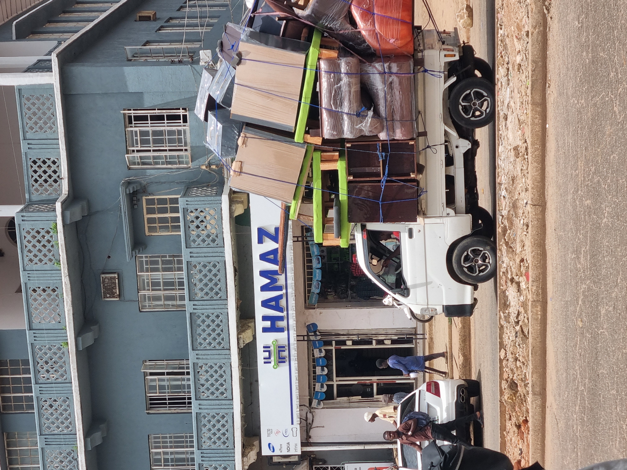 The image shows a white truck overloaded with furniture parked in front of a storefront with a large sign that reads "HAMAZ" vertically. The sign also features the names of phone brands, including "TECNO," "Samsung," and "Infinix," indicating a mobile phone store. Several people are present near the parked vehicles in front of the store. The building behind the store is a pale blue color with multiple windows and decorative screens. The location is likely in Jos, Nigeria, given the text on the store sign and the presence of local residents.