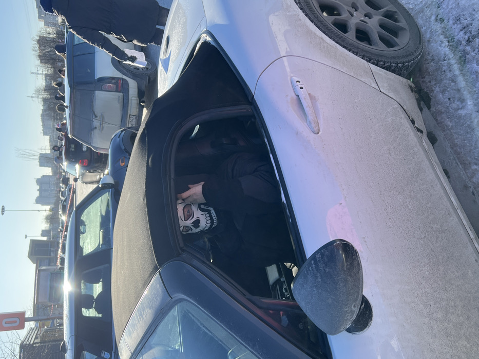 The image captures an outdoor scene on a clear, sunny day, likely in a large parking lot or open urban area in Saint Petersburg, Russia. The ground surface shows patches of dirty snow and slush.
In the foreground, a white Mazda MX-5 (ND generation) convertible is prominently featured, heavily soiled with road grime, indicating recent travel in adverse conditions. Its black soft-top roof is partially raised, and the driver's side window is lowered. A person occupies the driver's seat, dressed in a black puffer jacket and a white balaclava or mask adorned with a black skull pattern, covering the face except for the eyes. The individual's right hand is raised near their face.
In the mid-ground, a dark-colored Mini Cooper is parked directly behind the convertible. Further back, a soiled white Mini Clubman (first generation) is visible, displaying a Russian license plate "P263MT 178," which identifies its registration region as Saint Petersburg. To the right of the frame, a person wearing a dark winter jacket walks away from the camera, holding white papers in their left hand. The background includes numerous other parked vehicles and indistinct figures, indicating a populated area or gathering. Distant urban infrastructure, including a building with a large red "0" sign and residential high-rise buildings, completes the horizon.
