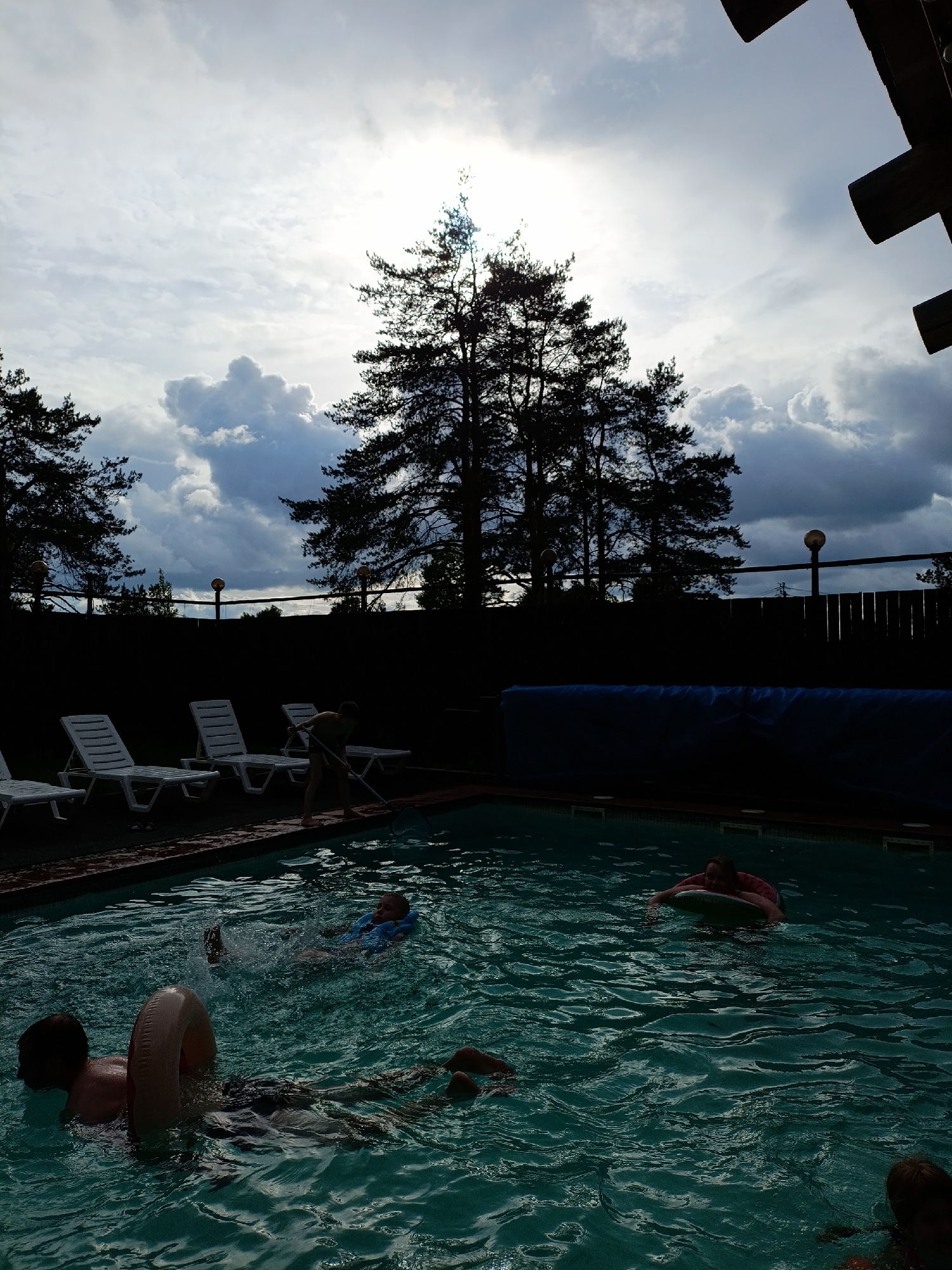 The image captures an outdoor swimming pool scene, likely at a dacha or resort in Chaikovsky, Russia, under an overcast sky. The overall lighting is backlit, silhouetting the surrounding elements and creating a moody atmosphere.
In the foreground and midground, several people are visible in the swimming pool. On the left, a man is partially submerged, wearing swim trunks and holding onto a beige inflatable ring. To his right, a child splashes in the water, wearing a blue life vest or buoyancy aid. Further back and to the right, another person is lying on a green inflatable float. The ripples and splashes in the water indicate active swimming and play.
Along the pool's edge, on the left, several white plastic lounge chairs are lined up. To the right of these chairs, a person, possibly a child or young adult, stands on the poolside, actively cleaning the water with a long-handled net.
The background features a dark wooden fence running horizontally. Above the fence, tall, dark pine trees dominate the upper half of the image, appearing as silhouettes against the bright, cloudy sky. Two spherical street lamps with orange glows are mounted on posts behind the pool, adding a touch of artificial light. In the top right corner, part of a rustic wooden structure is visible. The sky is largely covered with light and dark grey clouds, with some brighter areas suggesting the sun's position.
No visible text is present in the image. The scene depicts a leisurely summer activity, with people enjoying a swim despite the cloudy weather, while someone attends to pool maintenance.