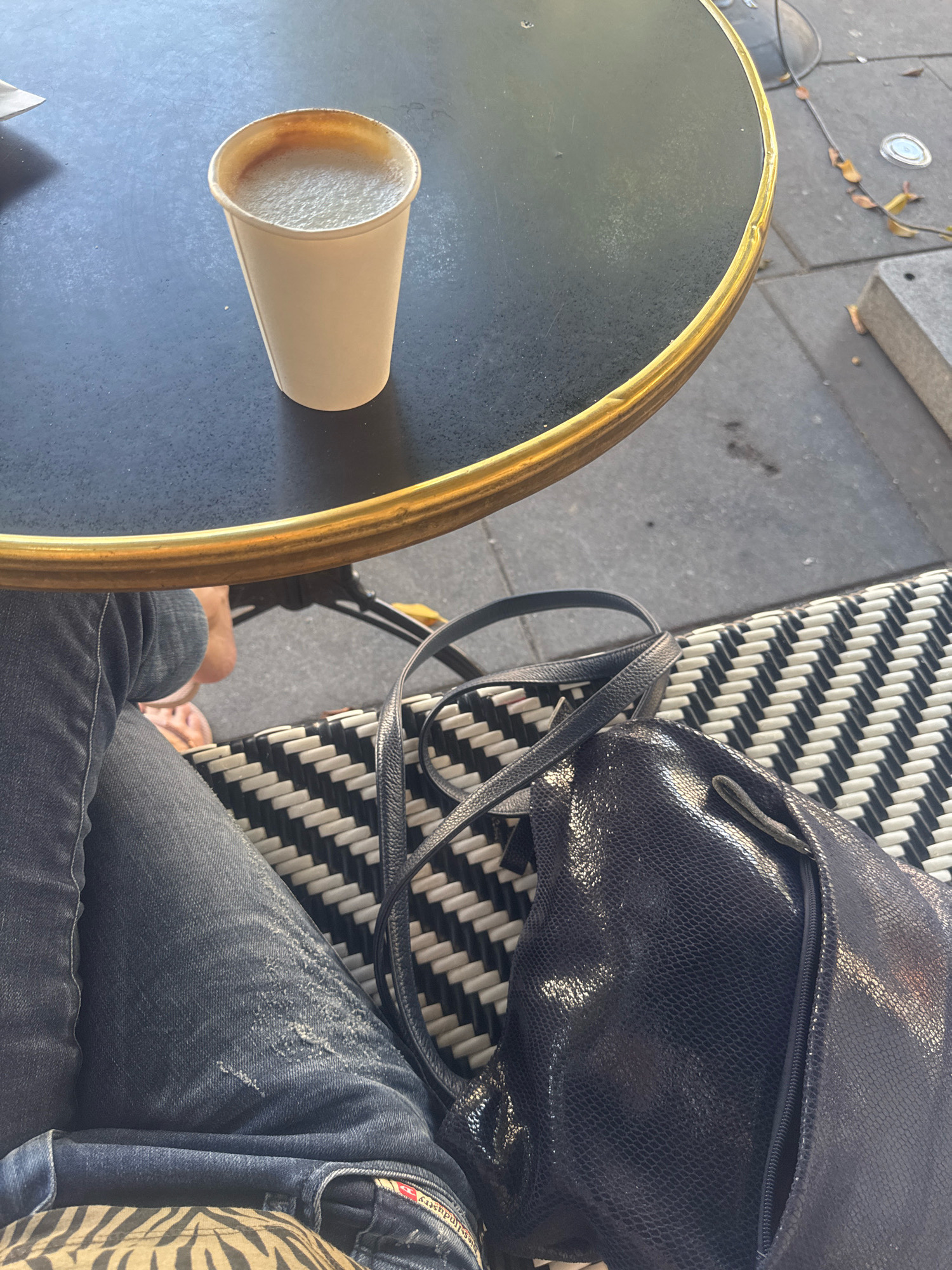 A close-up, slightly elevated medium shot shows a person sitting at an outdoor table in Ramat Gan, Israel. The table has a black surface and a gold rim. A white paper cup filled with a frothy beverage, likely coffee, rests on the table. The person is wearing blue jeans and is seated on a bench with a black and white woven pattern. A dark blue, textured handbag with a zipper is placed on the bench next to the person. The background shows a gray paved area with some fallen leaves and a small metal object. The lighting suggests it is daytime.