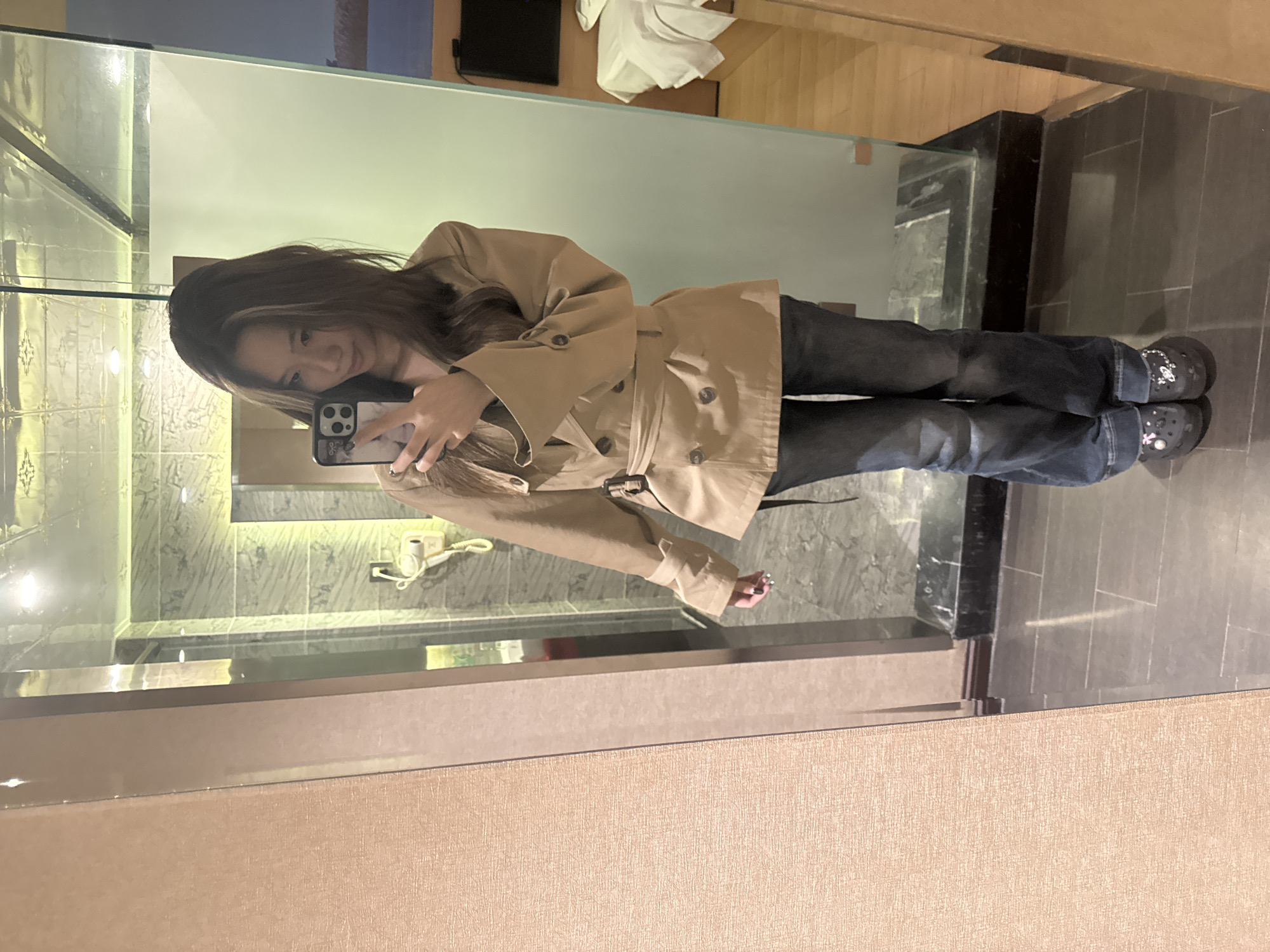 A young woman is captured in a mirror selfie, seen from the chest down. She holds a smartphone, partially obscuring her face, as she takes the picture. Her long, dark hair, with lighter highlights, falls around her shoulders. She wears a light brown or beige double-breasted trench coat, cinched at the waist with a matching belt, and dark blue jeans. On her feet are black clogs, possibly Crocs, adorned with small white or silver charms, including a flower and a star.
The setting appears to be a modern indoor space, likely a hotel room or a dressing area, in Ganzhou, China. The large mirror reflects a background featuring light-colored walls, some with subtle patterned tiling, and a section of wooden paneling. A dark television or monitor is mounted on a wall, and a white garment or towel hangs nearby. A beige corded telephone sits on a counter. The flooring is a mix of dark tiles and lighter wooden sections, separated by a dark, polished stone pillar. The lighting is artificial and warm. The woman has a calm, perhaps slightly smiling expression. No text is visible in the image.