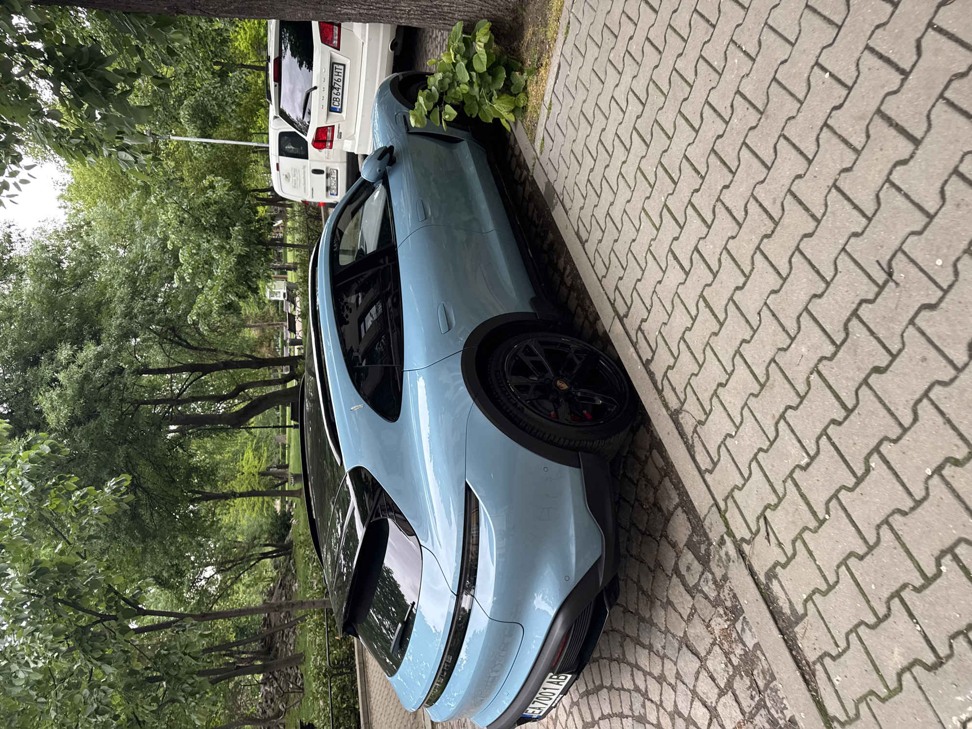 The image shows a light blue Porsche Taycan Cross Turismo parked on a sidewalk next to a street. The car is angled towards the curb and a tree. In the background, a white SUV and a white van are partially visible. The location is identifiable as Sofia, Bulgaria, due to the license plate "ЕН 7001 AB" on the Porsche. The sidewalk is paved with interlocking concrete pavers. Trees are present along the street, suggesting a residential or park area. The lighting indicates it is daytime. There are no people visible in the frame.