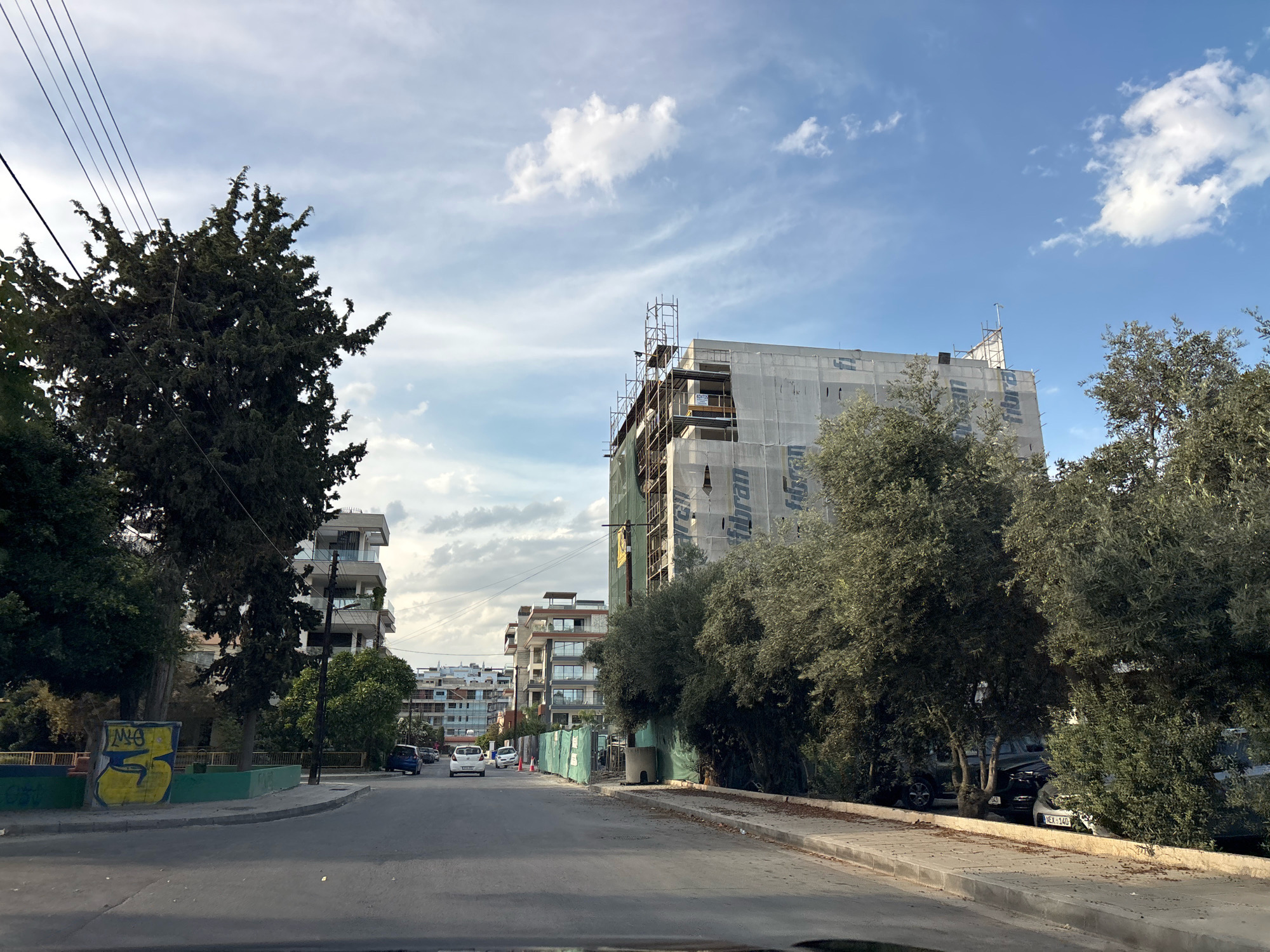 The image depicts a street scene in Germasogeia Municipality, Cyprus, with a focus on a multi-story building under construction. The building is covered in white protective sheeting and surrounded by scaffolding. Adjacent to it are other buildings, some appearing residential with balconies. Several mature trees are scattered throughout the scene, including a large, dark green conifer on the left and a cluster of olive trees to the right of the construction site. Power lines are visible running diagonally across the sky, which is partly cloudy. A paved road with painted lane markings leads into the distance, with a few cars visible, including a white car in the foreground and a dark blue car further down the street. A sidewalk runs along the right side of the road. Graffiti is present on a low wall to the left of the road. The overall impression is of an urban environment undergoing development.