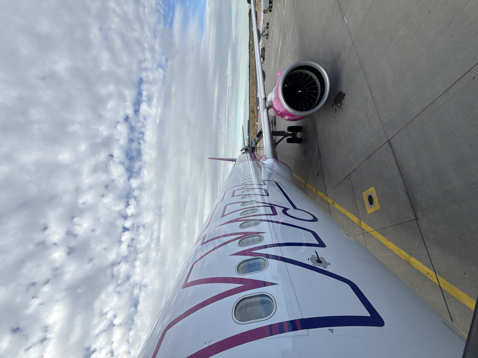 An exterior, eye-level shot shows a Wizz Air airplane parked on an airport tarmac under a partly cloudy sky. The fuselage of the plane, which is white with a large purple and pink Wizz Air logo, occupies the left and center portions of the frame. Several windows are visible along the fuselage. The right side of the plane features a bright pink engine nacelle with a silver fan. The tarmac is gray concrete with yellow and white lines. In the background, a mobile staircase is attached to the plane, and other airport equipment and a distant building are visible. The scene appears to be an airport in or near Künersberg, Germany, given the Wizz Air livery and common airport features.