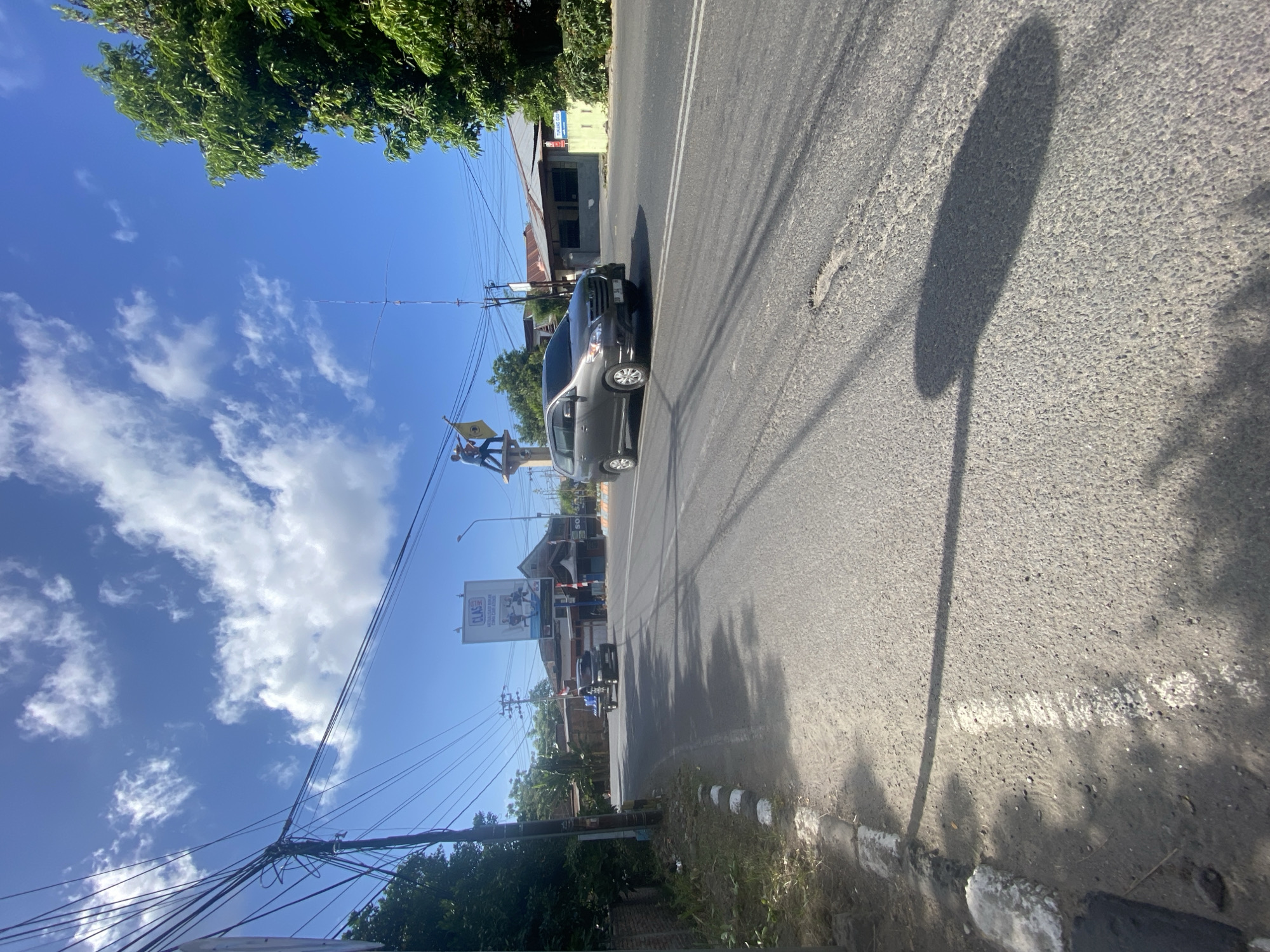 In Bentengnge, Indonesia, a grey minivan drives on a paved road under a bright blue sky with scattered white clouds. The road has two white lines marking lanes and a pothole is visible in the right lane. To the left of the road, a sidewalk with white and grey curbing runs alongside. A utility pole with numerous wires stands on the left side of the road. In the distance, a billboard with the word "CLAS" is visible above buildings. A statue of two people holding flags stands on a pedestal near the billboard. A smaller black vehicle is seen on the road in the distance. Lush green trees are present on the right side of the road and in the background. The sun casts shadows on the road, indicating a daytime scene.