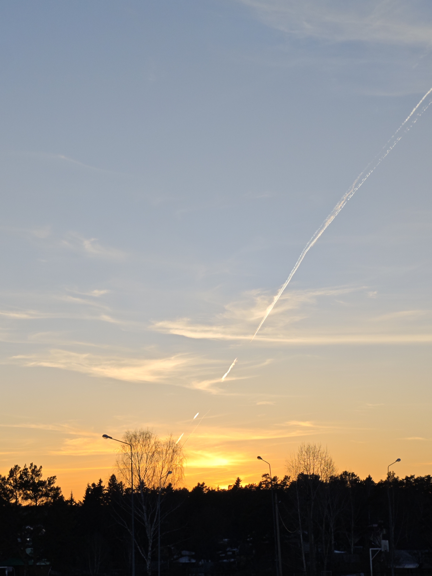 The image shows a sunset over the city of Chaikovsky, Russia. The sky transitions from orange near the horizon to light blue higher up, with contrails visible. The horizon is lined with a silhouette of trees. Street lamps are visible among the trees. In the foreground, there are some dark structures, possibly buildings.