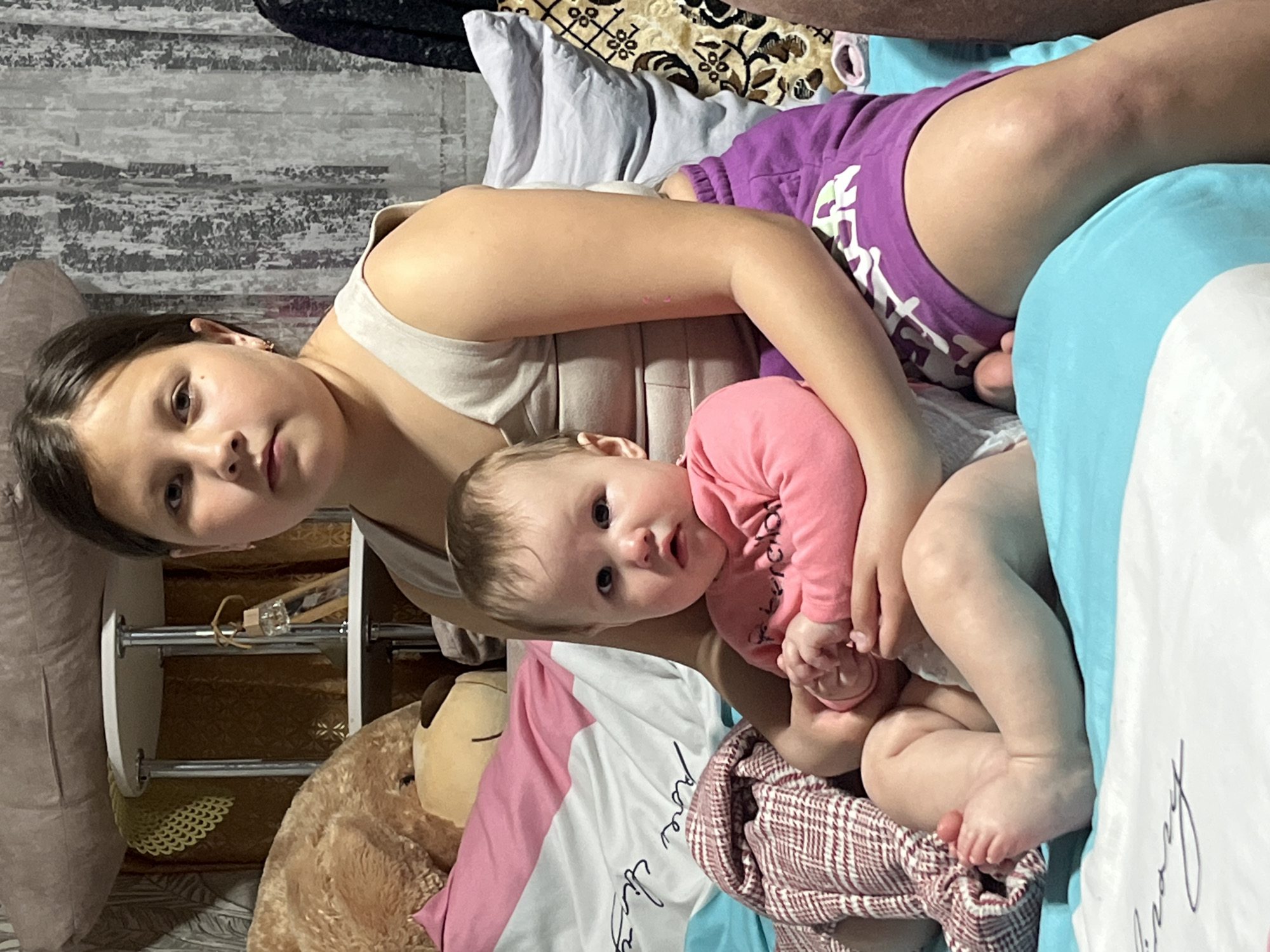 The image shows a young girl holding a baby in a room. The girl is fair-skinned with brown hair, wearing a light-colored tank top. The baby, also fair-skinned with light hair, is wearing a pink long-sleeved shirt. They are sitting on a bed with multiple pillows and blankets of varying colors and patterns (blue, pink, white with black writing, patterned browns). A stuffed teddy bear is nearby. Behind them is a small, white shelving unit and a wall with a gray textured pattern. The location is identified as Novopokrovskaya, Russia.