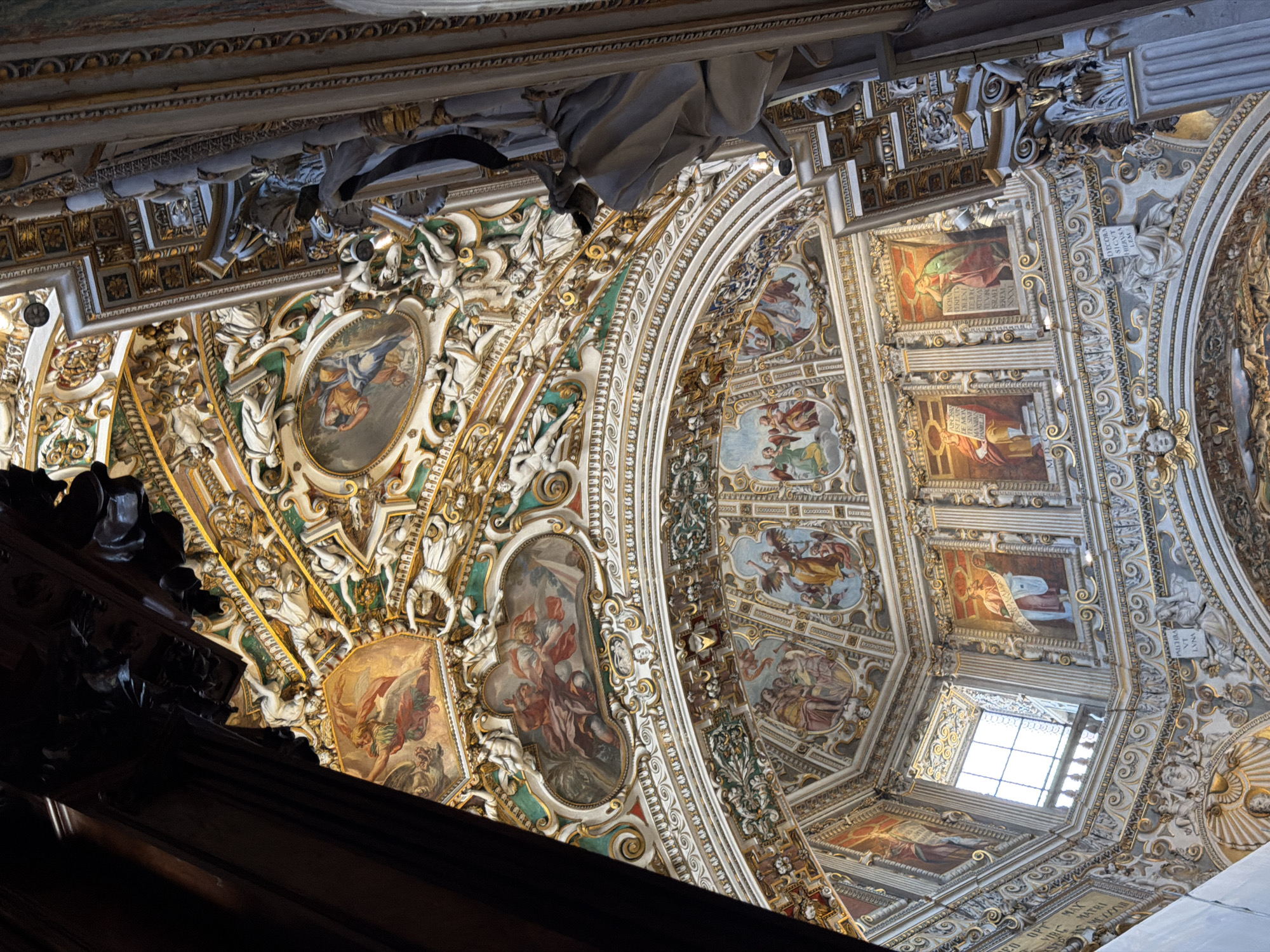 This is a close-up, upward-facing view of the ornate ceiling of the Church of San Giorgio Martire in Mozzo, Italy. The ceiling is a complex architectural and artistic composition featuring a dome with painted frescoes and intricate stucco work, all heavily adorned with gold accents.
The frescoes depict religious scenes with numerous figures in traditional attire, rendered in rich colors. These paintings are framed within oval and rectangular cartouches, surrounded by elaborate gilded stucco ornamentation, including cherubs, drapery, and architectural motifs. The stucco work itself is highly detailed, creating a sense of depth and movement.
The viewpoint suggests the photographer is standing within the nave of the church, looking up towards the crossing or a dome. A large dark wooden architectural element, possibly a beam or part of a choir loft, is visible in the foreground on the left, partially obscuring the ceiling. On the right side of the image, a large, light-colored sculpted figure, possibly a saint or angel, is positioned on a column, further emphasizing the grand scale and decorative richness of the interior. The overall impression is one of opulent religious artistry and architecture.