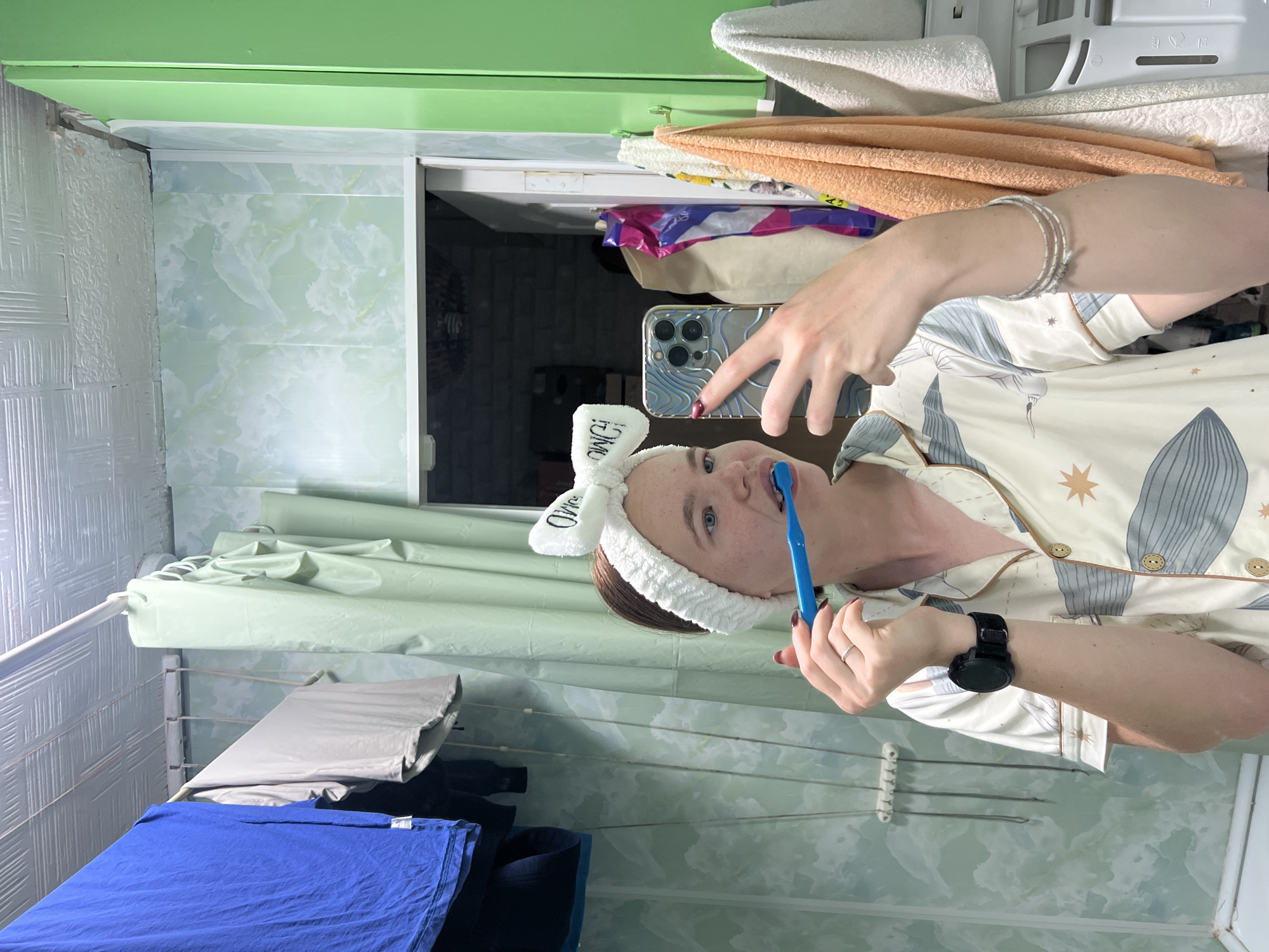 A young woman is brushing her teeth in a bathroom, reflected in a mirror. She is wearing a white fuzzy headband with the word "OW!" printed on it, and pajamas with a celestial pattern. She holds a blue toothbrush in her right hand and a smartphone in her left, using it to take a selfie. A black smartwatch is on her left wrist, and multiple bracelets are on her right wrist. The bathroom features light green marbled wallpaper and a white tiled ceiling. Clothes hang on a line in the background. Towels and other toiletries are visible on shelves to the right. The scene appears to be in a residential setting. The image does not provide any specific information to pinpoint its location within the city of Chaikovsky, Russia.