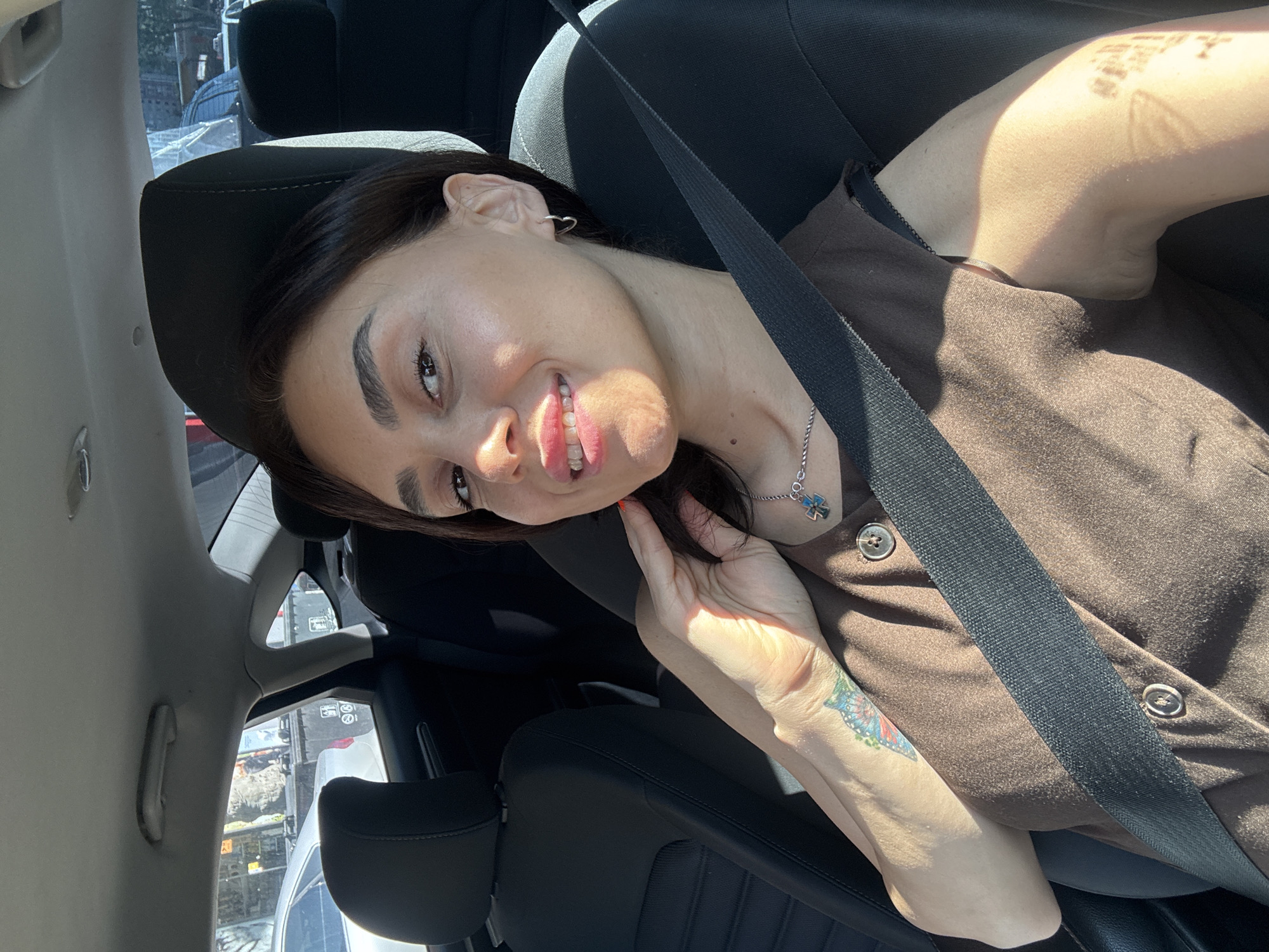 The image shows a woman sitting in the passenger seat of a car. She is wearing a brown top and a seatbelt. She has a tattoo of a butterfly on her left arm. The car appears to be parked on a street with other vehicles and buildings visible in the background. The lighting suggests it is daytime. It is not possible to determine the specific location in Tbilisi, Georgia based on the visual information provided.