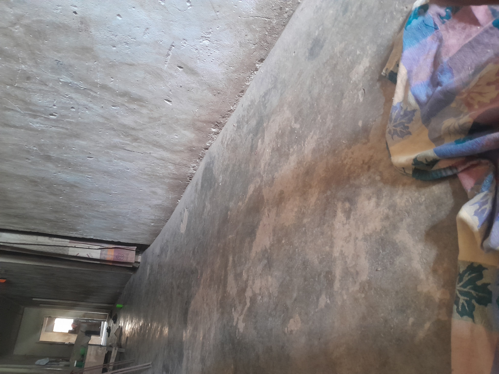 This image depicts a dimly lit concrete corridor within a building in Ifo, Nigeria, viewed from a low angle. The floor is unpolished concrete, exhibiting mottled patterns of gray and brown with some reflective areas. The walls are rough, unpainted concrete or plaster, with the right wall displaying significant texture and minor deterioration along its base. In the immediate foreground, a folded or bunched fabric, patterned with blue, purple, beige, and green floral or geometric designs, rests on the floor. Towards the midground on the left, a wooden structure, possibly a table or workbench, is visible, accompanied by several thin wooden supports leaning vertically. A small, bright green plastic cup is on the floor near the left wall, and two dark-colored sandals are positioned on the floor closer to an open doorway. Through this open, brightly illuminated doorway at the far end of the corridor, a partially obscured individual is discernible standing, with other indistinct objects visible in the background room. No direct actions or interactions between individuals are clearly depicted.