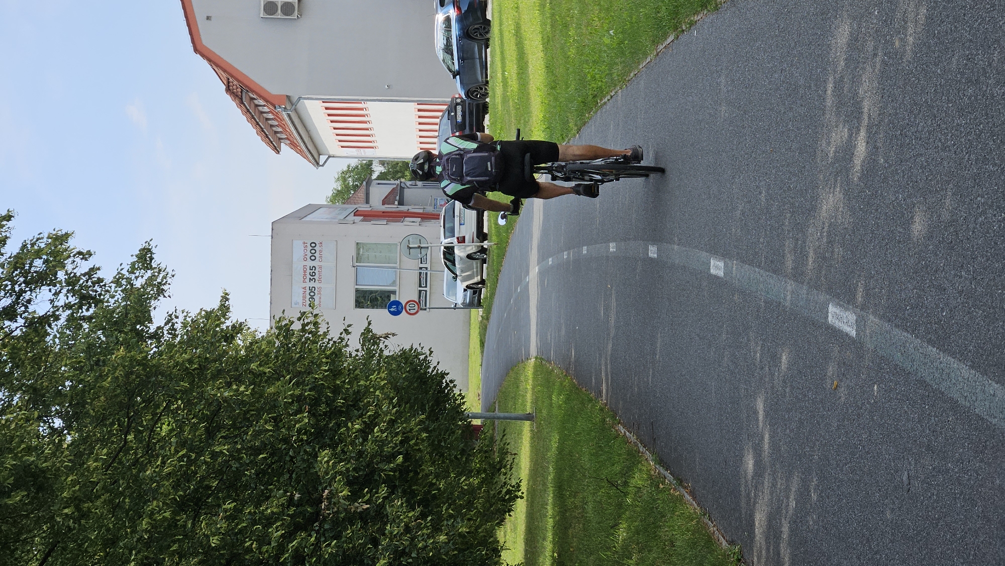 A person is cycling on a paved bike path in Prešov, Slovakia. The path is bordered by green grass and a large tree on the left, and more grass on the right. In the background, there are buildings, including a modern white building with a sign that reads "ZUBNA POHOTOVOSŤ" and "365 dental care.sk", and a building with a red-tiled roof and red window frames. Several cars are parked near the buildings. The sky is blue with some clouds. The cyclist is wearing a helmet and a backpack.