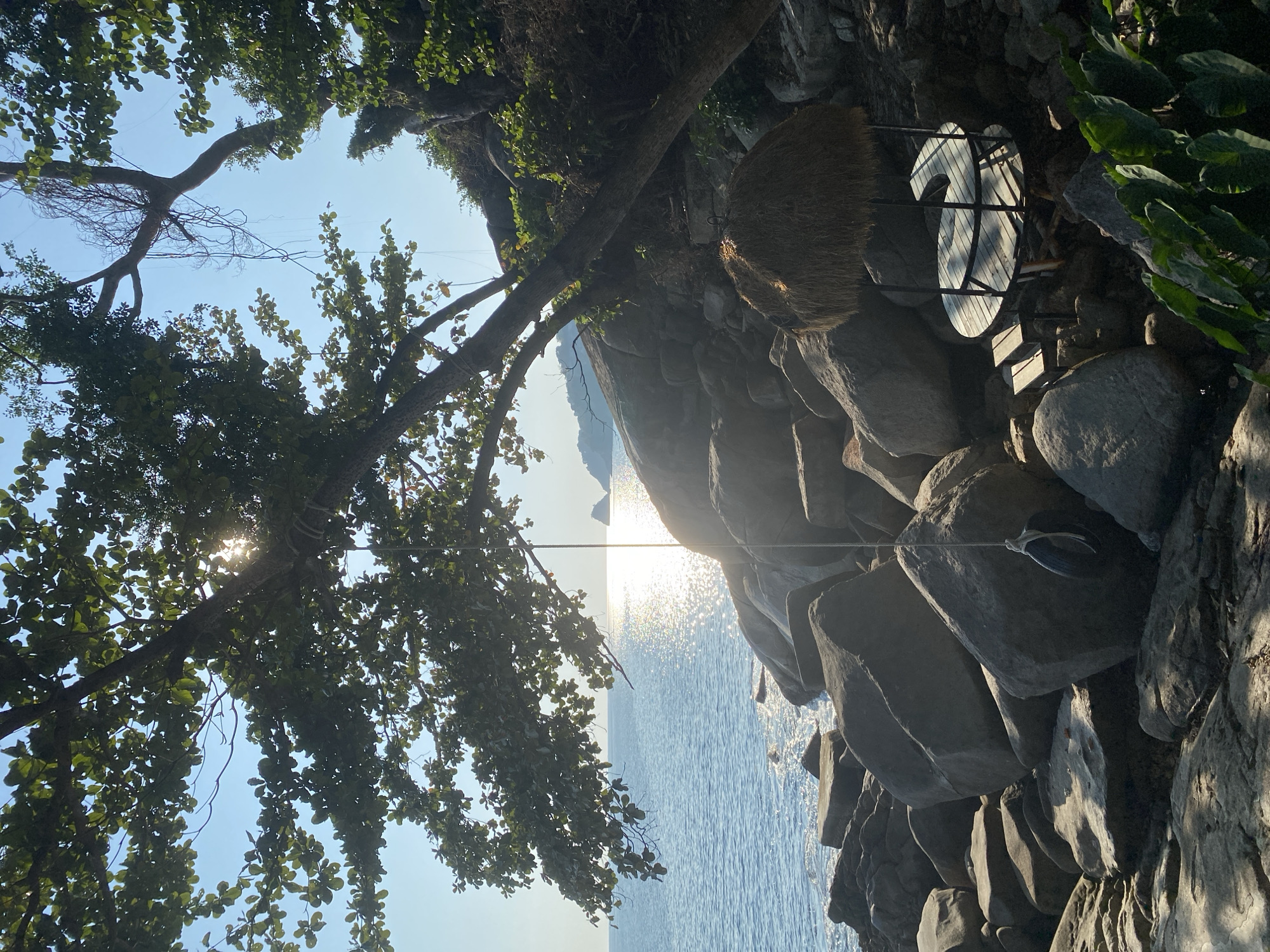 The image depicts a view from a rocky cliffside in Patong, Thailand. The location appears to be near the coast, overlooking the ocean. The sun reflects brightly on the water. A tree with dense foliage extends over the cliff. A sign with a number "2" on it is affixed to the rocks, along with a woven, circular decoration. The rock formation is composed of large, uneven boulders.