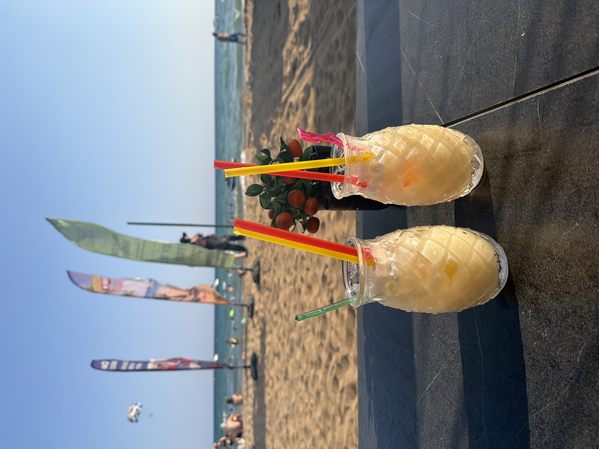 Two pineapple-shaped glasses filled with a light-colored beverage and ice, each with two straws, are placed on a dark, textured surface. One glass is closer to the viewer than the other. In the background, a sandy beach stretches towards a calm sea under a clear blue sky. Several colorful flags are planted on the beach, and a person is parasailing in the distance. People are visible on the beach, some sitting and others walking towards the water. The scene appears to be a waterfront bar or restaurant in Manavgat, Turkey.