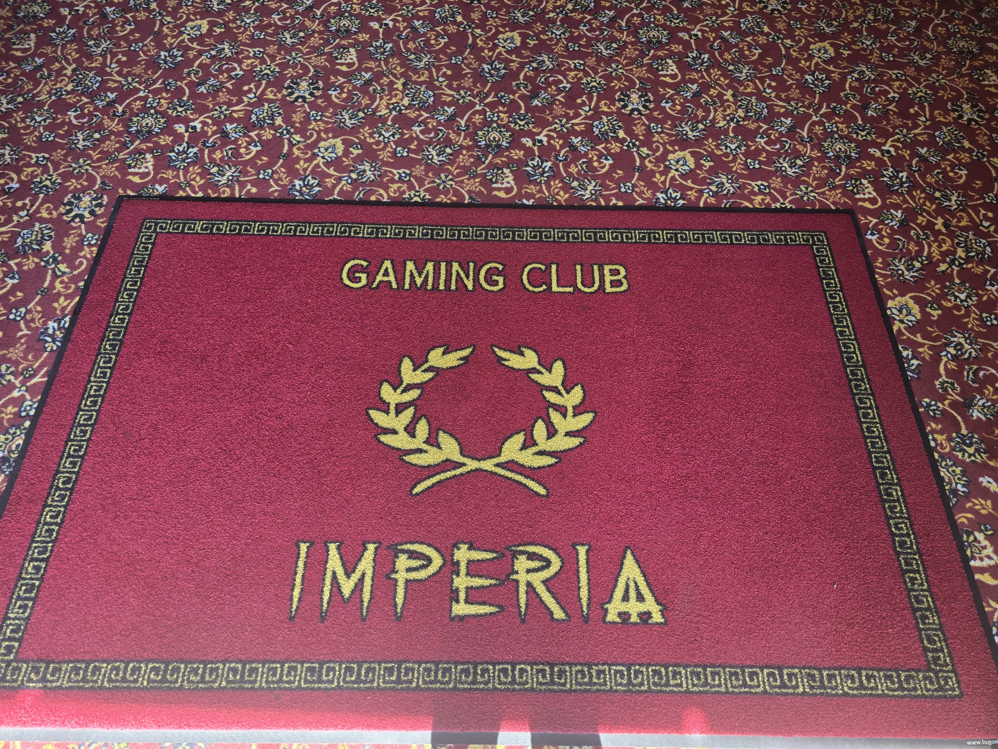 The image shows a close-up of a red doormat with gold lettering and a laurel wreath design. The doormat is placed on a patterned carpet with a floral design in shades of red, yellow, and blue. The doormat has a decorative border with a Greek key pattern. The lettering on the doormat reads "IMPERIA" vertically on the left side and "GAMING CLUB" vertically on the right side. There is no visible indication of Malko Tarnovo, Bulgaria in the image. The scene is an indoor setting, likely an entranceway or a gaming establishment.