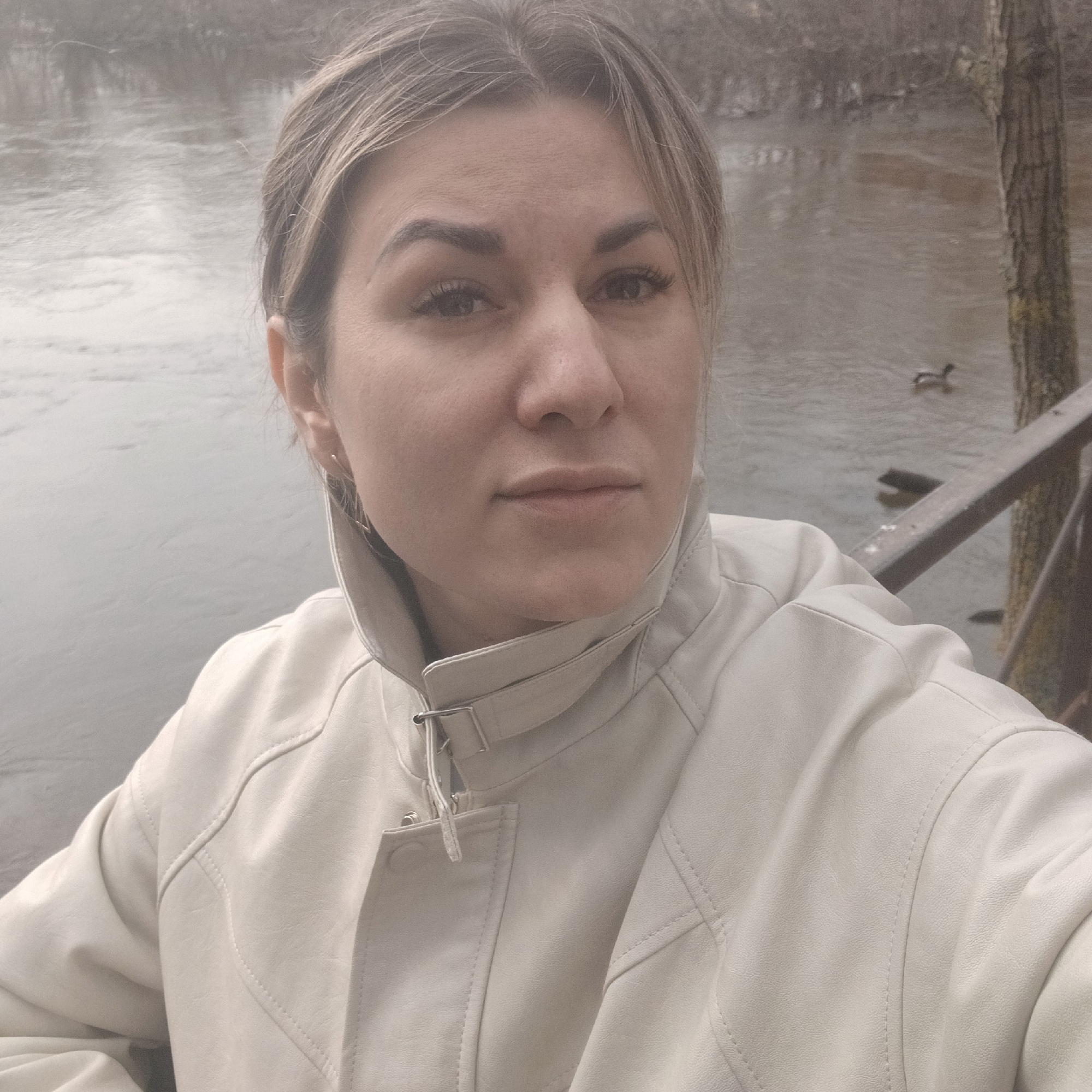 A woman stands in Izhevsk, Russia, wearing a light-colored jacket. She is near a body of water with trees and a railing visible in the background. A duck is visible in the water.