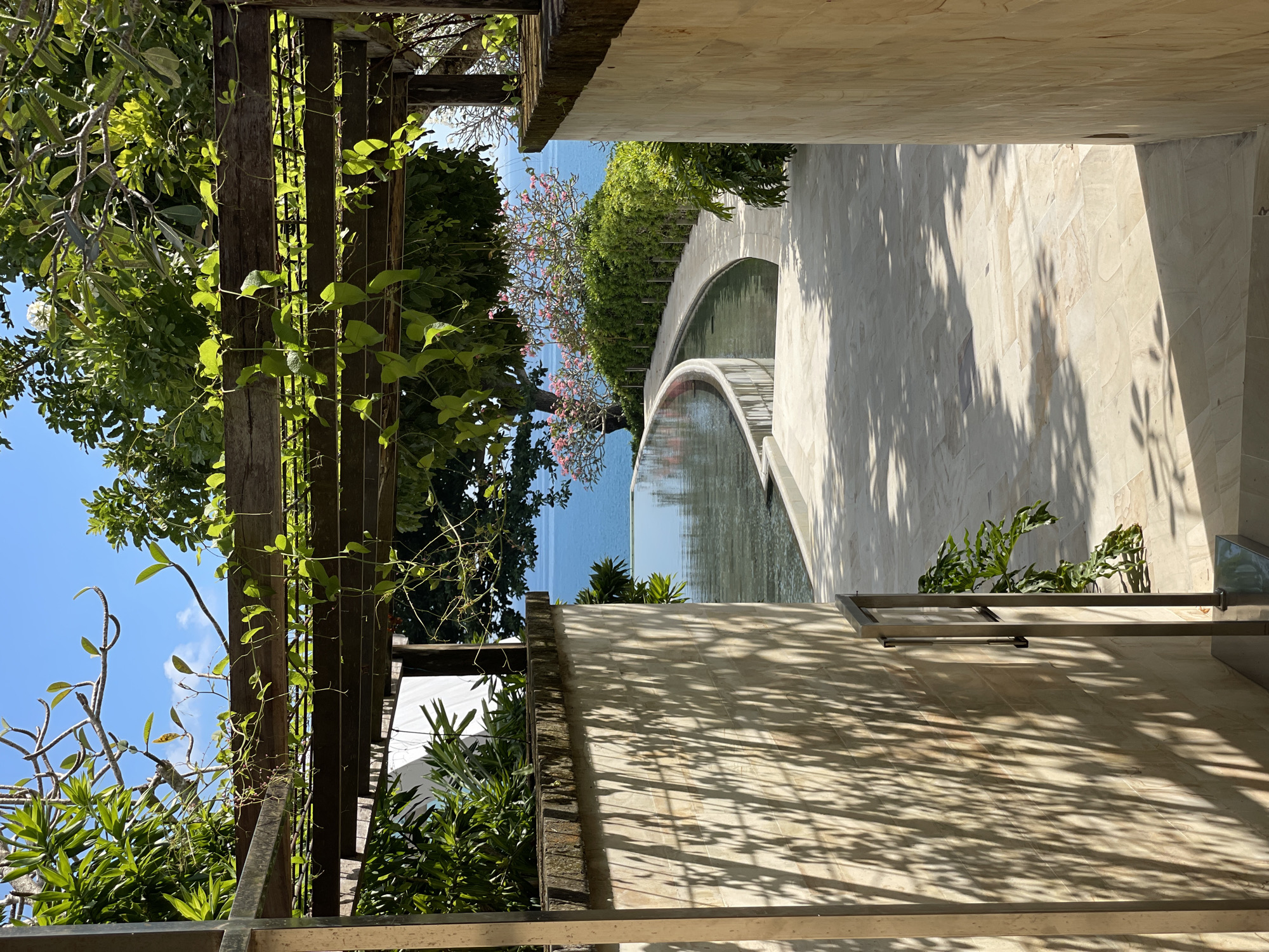Captured from an elevated perspective, the photograph reveals an outdoor scene in an unknown city. A weathered wooden pergola, partially covered in climbing vines, stands on the left side of the frame, juxtaposed against a vivid blue sky dotted with clouds. The pergola casts intricate shadows on the wall.
Beyond the pergola, a view of the ocean is framed by lush greenery, including a tree adorned with pink flowers. A marble bridge arches over a pool of water, reflecting the sky. The right side of the image features a light-colored stone wall, textured with geometric patterns and covered in leafy shadows.
There are no people visible in the image. The focus is on the architectural elements, the natural vegetation, and the tranquil water.