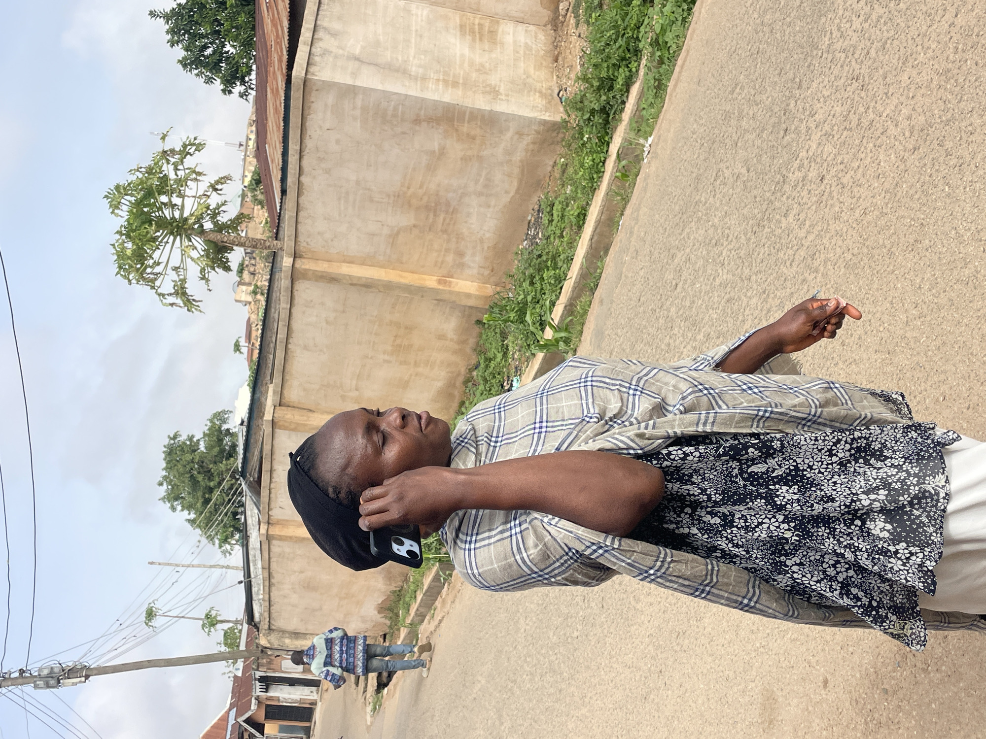 A person stands on a paved street in Jos, Nigeria, holding a black phone to their ear with their left hand. The person is wearing a black head covering, a plaid button-down shirt, a floral print top, and white pants. Another person, wearing a blue and white patterned shirt, is walking away in the background. Utility poles and wires are visible on the left side of the frame. The background includes cement buildings with tin roofs, trees, and other greenery. The lighting indicates it is daytime.
