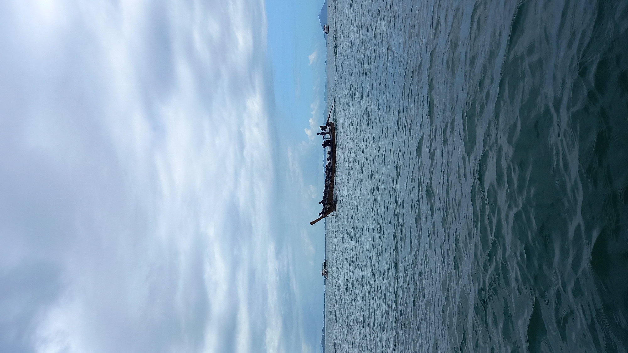 A longtail boat carrying approximately 20 people is traveling on the water in Ao Nam Mao, Thailand. The sea is slightly choppy, and the sky above has scattered clouds. A distant mountain can be seen on the horizon.