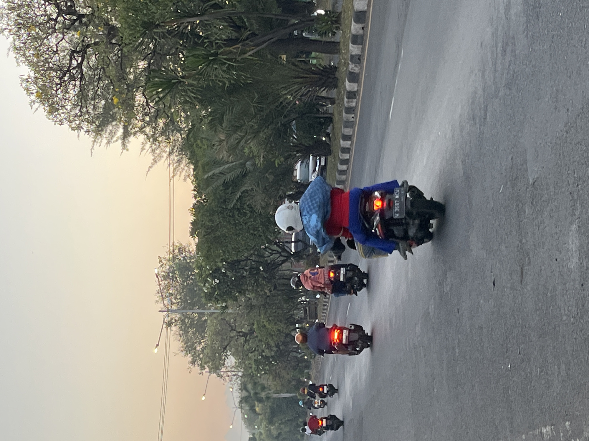 The image captures a street scene in Ngaban, Indonesia, likely during the early morning or late afternoon given the soft, warm lighting. Several individuals are riding motorcycles on a paved road. In the foreground, a person wearing a white helmet and a red shirt is riding a dark-colored scooter, carrying a blue object wrapped in fabric over their shoulders. Behind them, other motorcyclists are visible, some with passengers. The road is bordered by a curb with black and white markings and a sidewalk with lush green trees and vegetation. In the background, more trees and some buildings or structures are present, with a light-colored sky indicating the time of day. The overall impression is of daily life and commuting in a tropical urban environment.