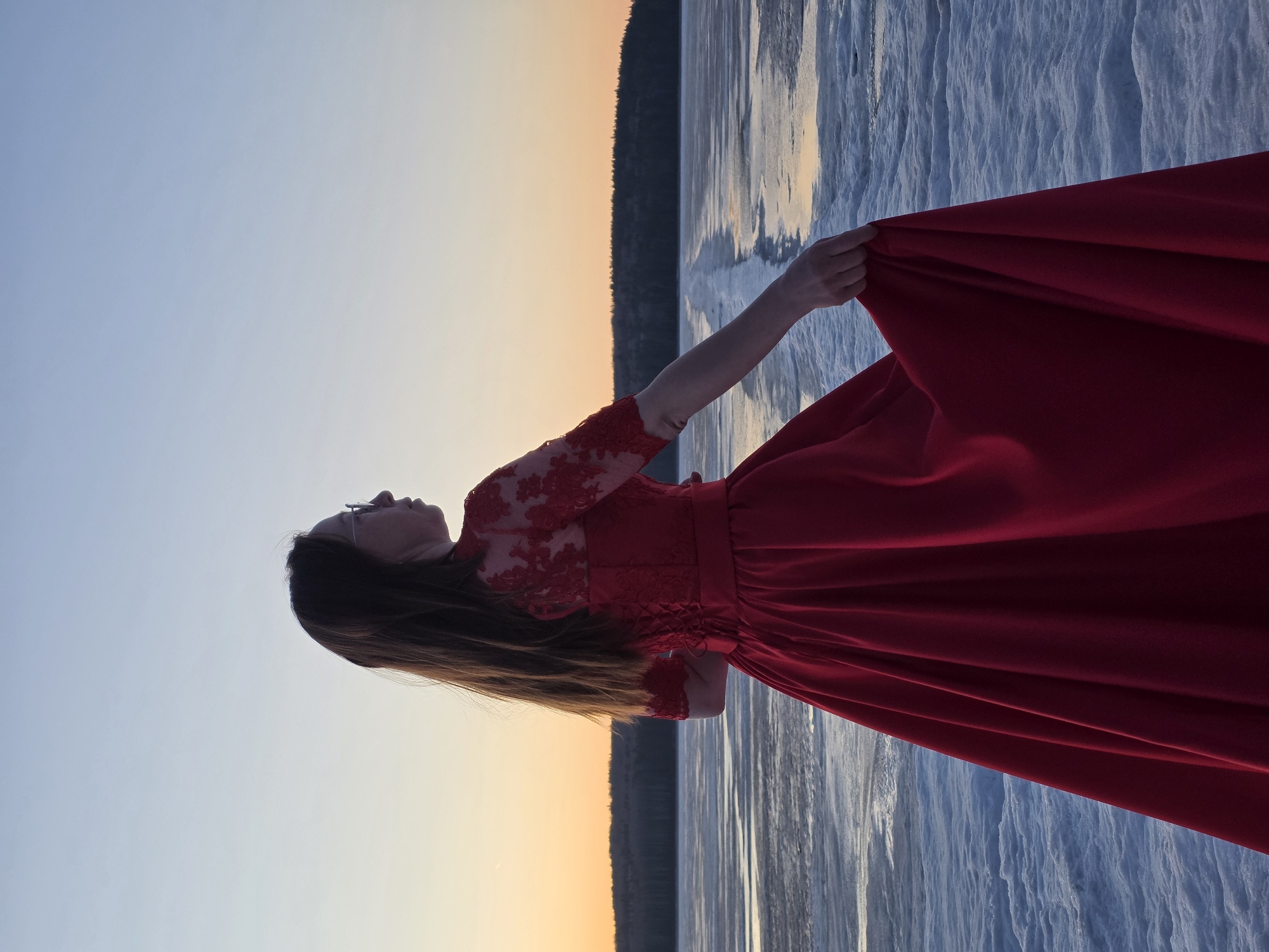 A woman wearing a red, long-sleeved dress stands on a snow-covered surface, possibly a frozen body of water, near Chaikovsky, Russia. She holds the hem of her dress in her right hand. The background consists of a gradient sky transitioning from blue to orange and a dark line of trees on the horizon. The ground has a textured, icy appearance.
