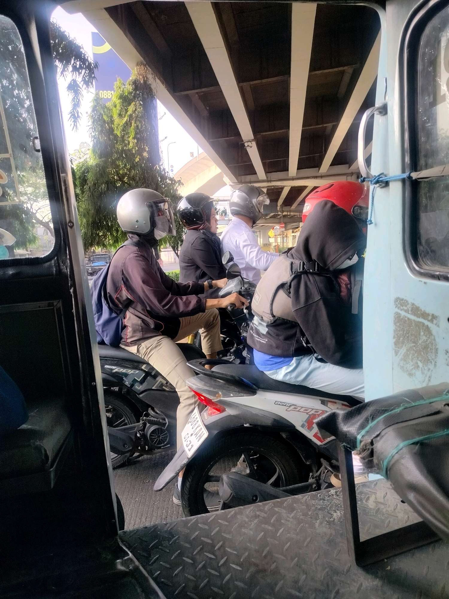 The image depicts a street scene in East Jakarta, Indonesia, likely during daytime. In the foreground, the interior of a vehicle, possibly a tricycle or a similar mode of transport, is visible. The interior features a dark metal floor with a diamond plate pattern. A black padded seat is also partially visible.
Outside the vehicle, several individuals on motorcycles are present. The first motorcycle is ridden by a person wearing a silver helmet and a dark jacket over khaki pants, with a blue backpack on their back. They are wearing a face mask. Behind them, another individual wearing a black helmet and a dark top is also on a motorcycle. Further back, a person in a white shirt is visible on a motorcycle, and another person wearing a red helmet and a black hooded jacket, along with a beige vest, is seated on a motorcycle behind them. The motorcycles appear to be common models found in the region.
The background shows a dense canopy of trees and vegetation, suggesting an area with greenery. Above the scene, an elevated road or bridge structure is visible, casting shadows on the vehicles below. A large billboard with text, including "0883" and some graphic elements, is partially obscured by trees and the bridge. The overall environment suggests a traffic situation on a city street, possibly near an overpass.