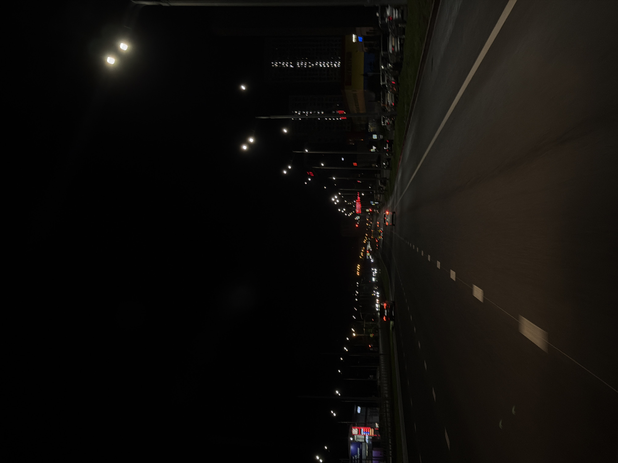 A nighttime, point-of-view shot captures a highway in an urban environment, illuminated by streetlights and the lights of passing vehicles and buildings. The road, with visible white lane markings, stretches into the distance, curving slightly to the right. On the left side of the highway, a row of streetlights stands, interspersed with commercial buildings and signage. On the right, parked cars line the roadside, and further down, taller buildings are visible, some lit up. The sky is dark, indicating late evening or early morning. The perspective suggests the viewer is in a moving vehicle, capturing the dynamic nature of a highway at night.