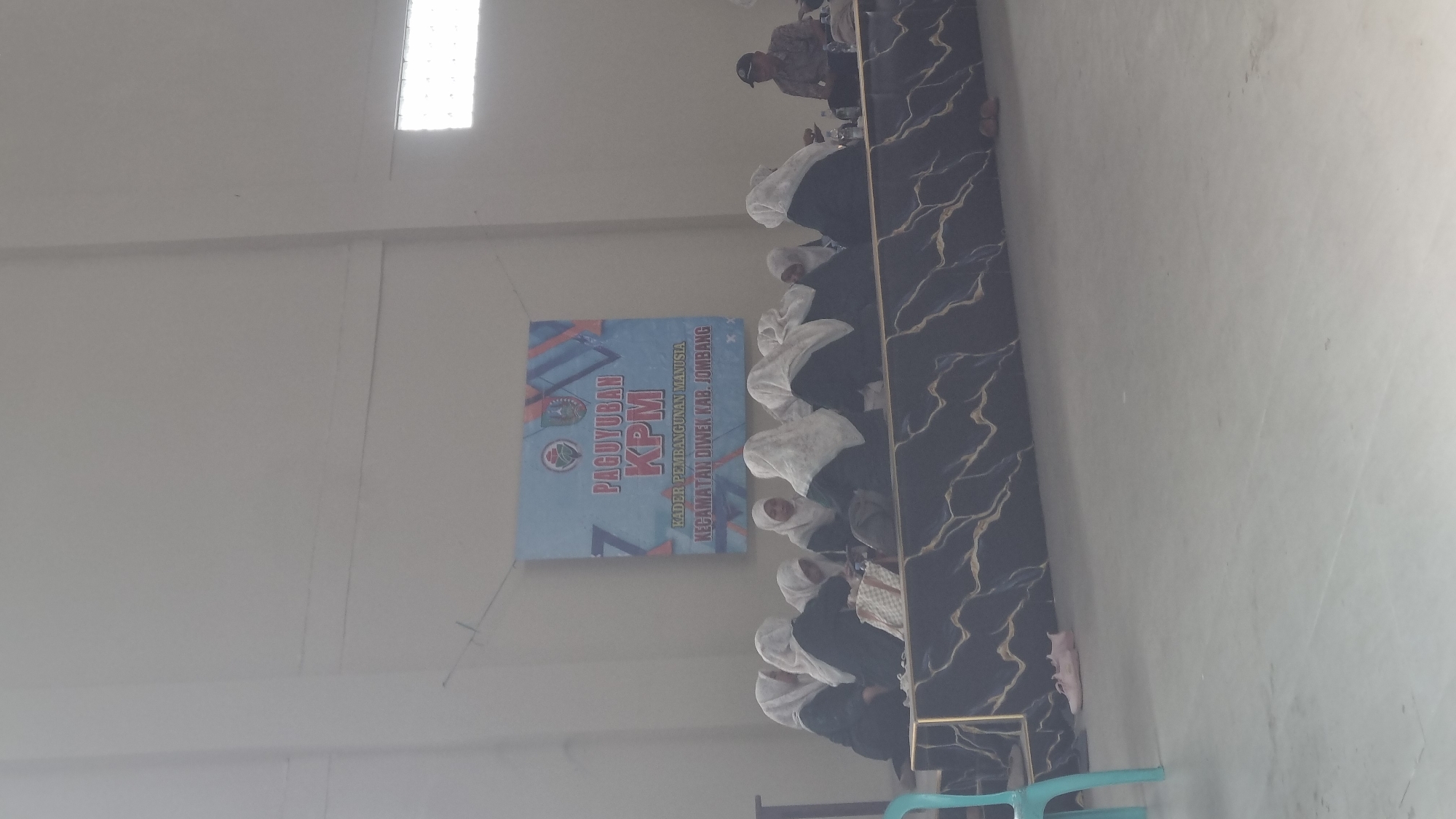 A group of people, predominantly women wearing hijabs, are seated in a room with a light-colored wall and floor, suggesting an indoor gathering space. A banner in the background displays "PAGUYUBAN KPM" and further text indicating "KADER PEMBANGUNAN MANUSIA KECAMATAN DIWEK KAB. JOMBANG," identifying the location as Diwek District, Jombang Regency, Indonesia. The participants are facing a table covered with a dark, marble-patterned fabric. Some individuals are looking down, possibly at phones or materials on the table. On the floor, a pair of light-colored sneakers is visible. To the right of the frame, a person wearing a cap and a patterned shirt is seated, appearing to observe the gathering. The overall ambiance suggests a meeting or training session.