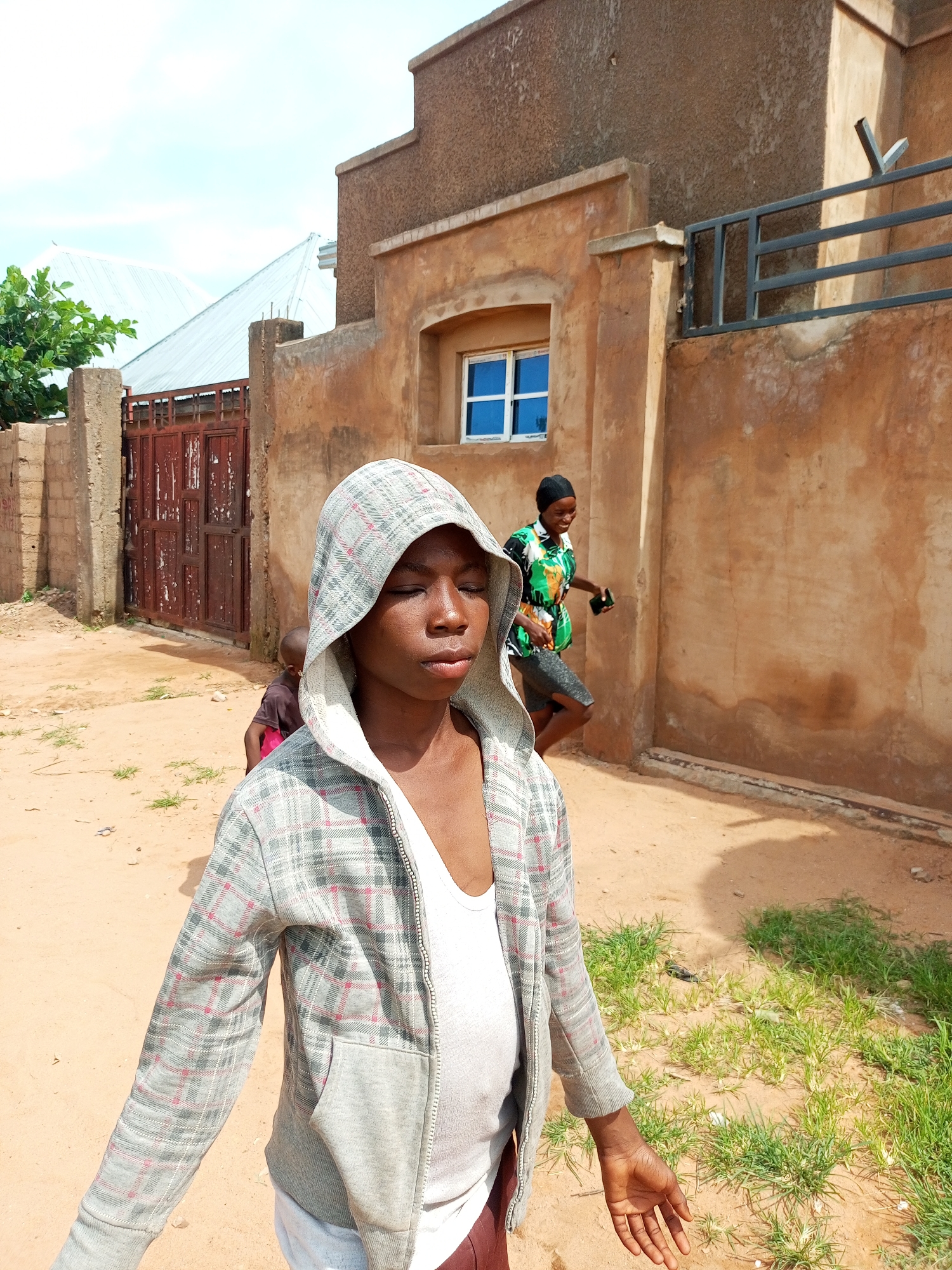 In the city of Tempure, Nigeria, the scene depicts an outdoor environment with a focus on individuals and buildings. A young person in the foreground wears a plaid hooded jacket over a white undershirt, with their eyes closed and head tilted slightly. Behind them, another individual in a colorful green and black patterned shirt and dark shorts is walking away, holding a dark object. Further in the background, a red metal gate with vertical bars is visible, adjacent to a building with a rough, textured brown facade and a blue-paned window. The ground is sandy, with patches of green grass growing in the area. A light-colored roof of another building can be seen in the upper left portion of the image.