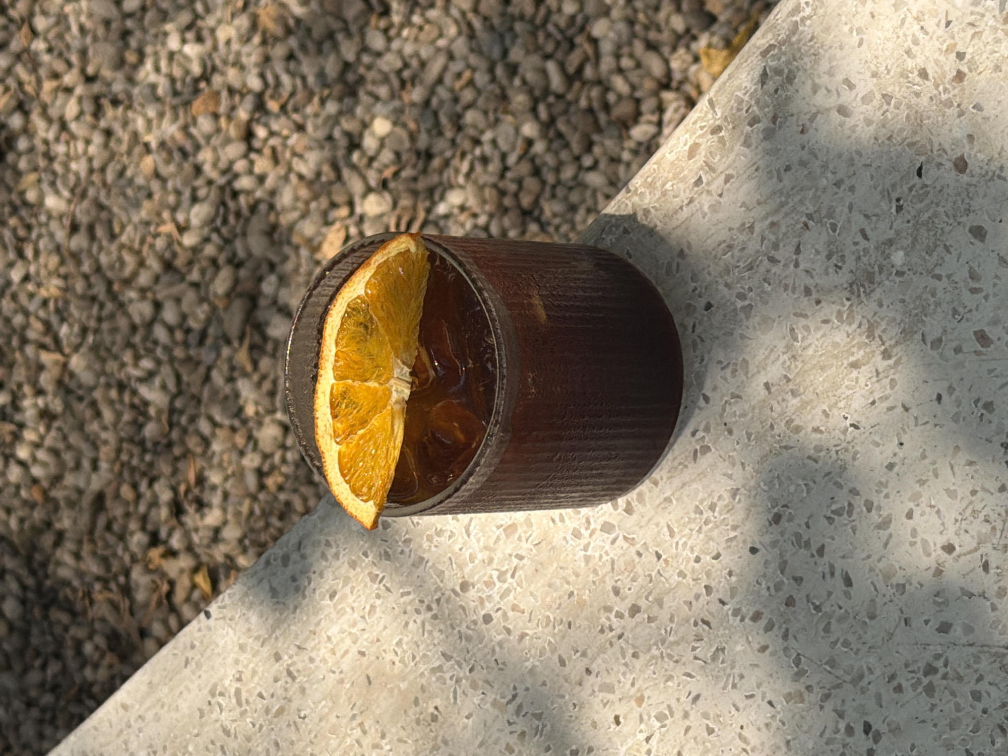 A top-down, eye-level, medium shot shows a dark glass containing a drink, garnished with a dried orange slice, positioned on a terrazzo surface. The glass is located at the junction where the terrazzo meets a surface covered in small pebbles. The terrazzo is a light color with darker speckles, and sunlight casts shadows across it. The glass is dark brown, cylindrical, and appears to hold a beverage with ice cubes, visible beneath the orange slice. The orange slice is positioned on the rim of the glass, with part of the slice submerged in the drink. The pebble-covered surface is predominantly gray with variations in tone due to light and shadow. The scene is outdoors and bathed in natural light, typical of Đà Nẵng, Vietnam, where outdoor cafes are common. No people are visible in the frame.