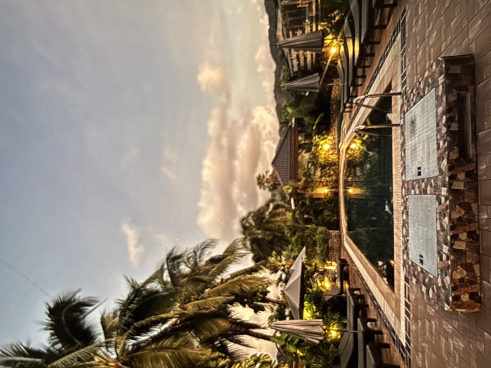 In the city of Chalong, Thailand, a rectangular swimming pool is the central focus of the scene. The pool's dark water reflects the twilight sky, which is a blend of pale blue and soft pink, with scattered, wispy clouds. Palm trees with dense fronds stand to the left, their leaves partially obscuring the sky and framing the pool area. Several lounge chairs are positioned along the poolside, some with closed umbrellas overhead. Yellow-toned lighting illuminates the foliage and the pool's edge, creating a warm ambiance. In the background, a gazebo-like structure with a dark roof is visible, surrounded by lush greenery and more palm trees. The pool deck is paved with brick-like tiles arranged in a patterned fashion. A small, tiled structure with a metal grate on top is situated in the foreground. The overall setting suggests a tranquil resort or hotel environment at dusk.