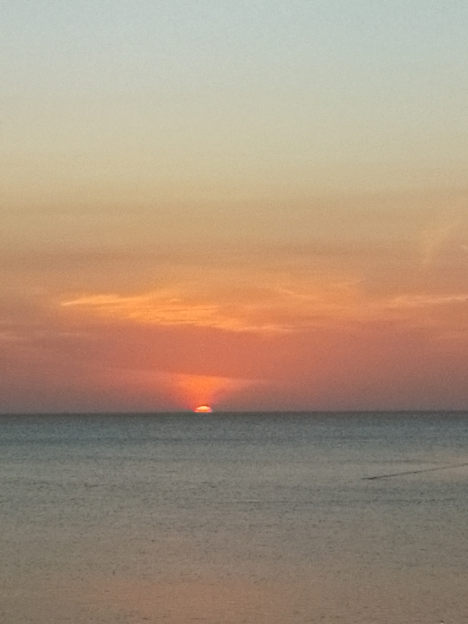 This image captures a breathtaking sunset over the ocean in Juan Griego, Venezuela. The scene is dominated by a clear horizon line separating the vast sky from the calm water.
The sun, a vibrant and intense orange-red, is prominently featured, appearing as a glowing semicircle partially submerged below the horizon, signifying the final moments of daylight. The sky above is a magnificent gradient of colors, transitioning from a soft, muted blue-grey at the top, through warm yellows and oranges, to rich reds and deep oranges closer to the setting sun. Wispy, elongated clouds are visible in the mid-sky, catching the golden light and enhancing the dramatic effect of the sunset.
The water surface below is relatively calm, reflecting the fading light in shades of deep blue-grey, with hints of purplish-brown towards the bottom of the frame. Subtle ripples are visible on the water, and a faint, dark line, possibly a distant boat or its wake, can be discerned towards the right side of the image.
The time of day is clearly late evening, during sunset. The weather appears clear and calm, contributing to the serene and peaceful atmosphere of the scene. There are no people or discernible text in the image. The overall impression is one of natural beauty and tranquility as the day concludes.