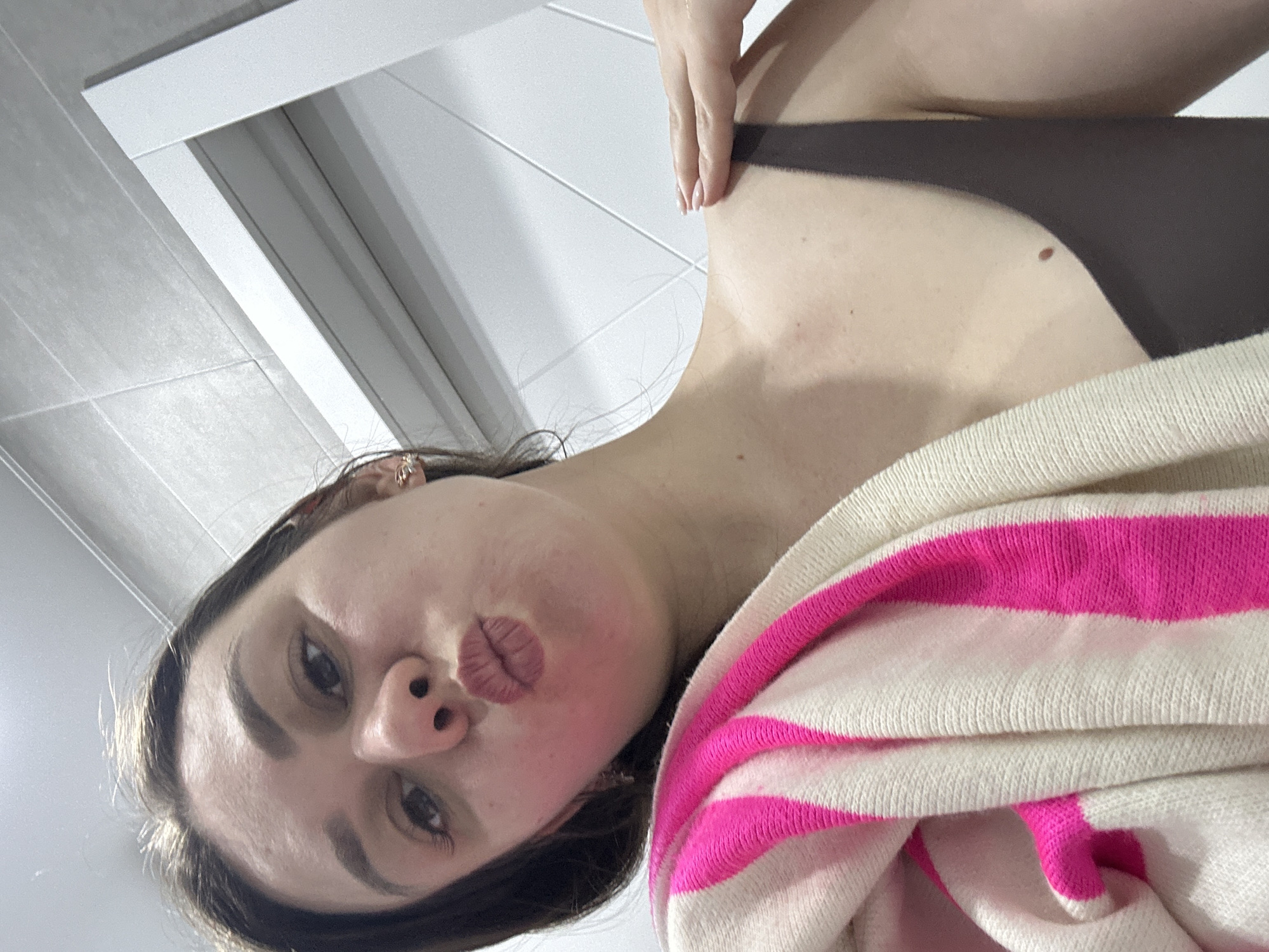 A woman is taking a selfie. She is wearing a brown top and a white sweater with pink stripes. She is puckering her lips. The location appears to be a bathroom. The scene appears to be indoors. There is no indication of the city of Chaikovsky, Russia.