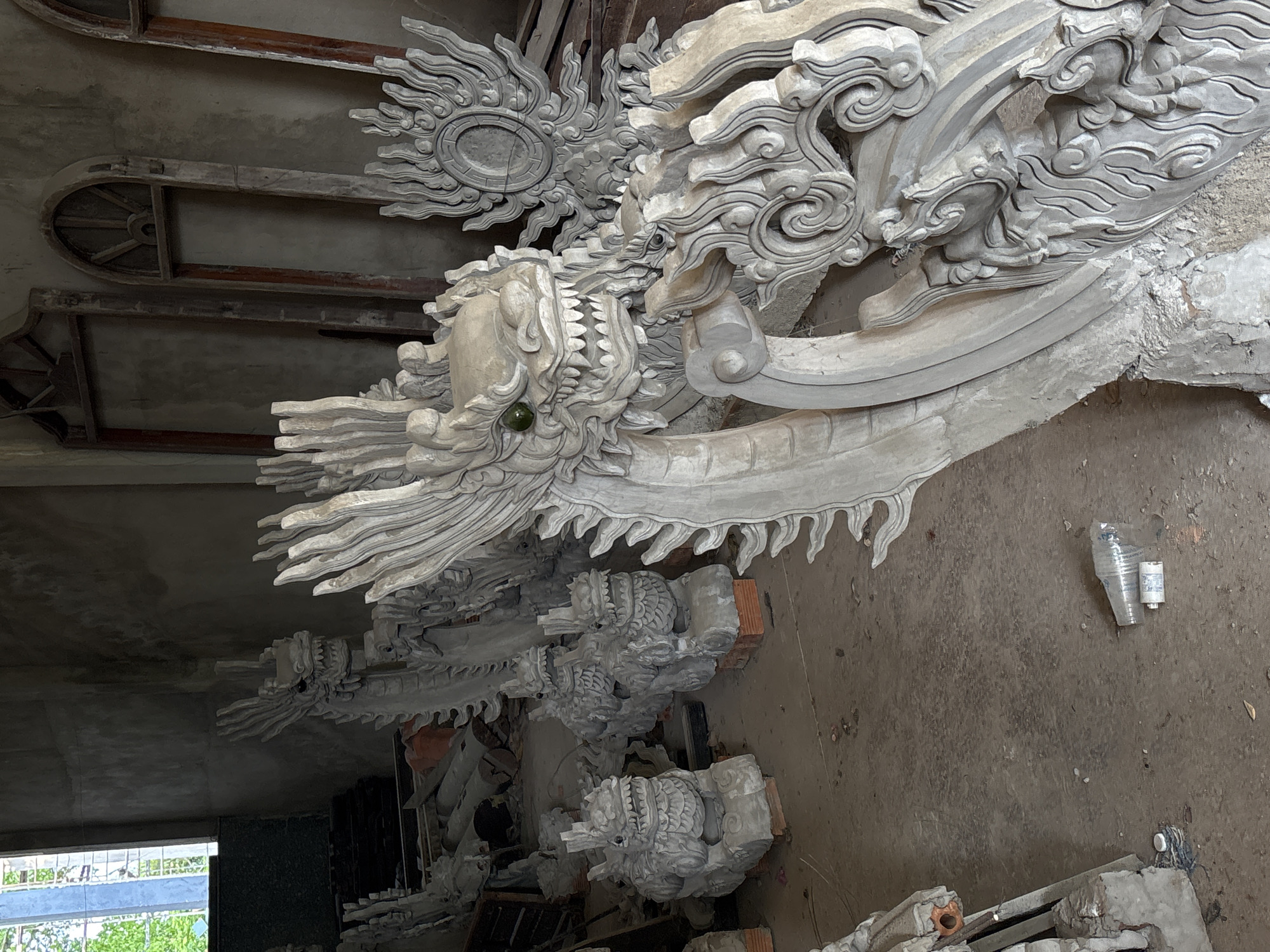 The image shows unfinished concrete sculptures of dragons and other mythical creatures in what appears to be a workshop or storage area. The sculptures are in various stages of completion, with some showing intricate details and others appearing more rudimentary. In the background, there are unfinished wooden window frames leaning against a wall. The floor is concrete and dusty, with some debris scattered around, including a plastic bottle and a small cylindrical object. The lighting suggests an indoor space, possibly with natural light coming from a window visible in the upper left corner. The style of the sculptures and the architectural elements in the background might suggest a location in Vietnam, potentially related to temple or pagoda construction.
