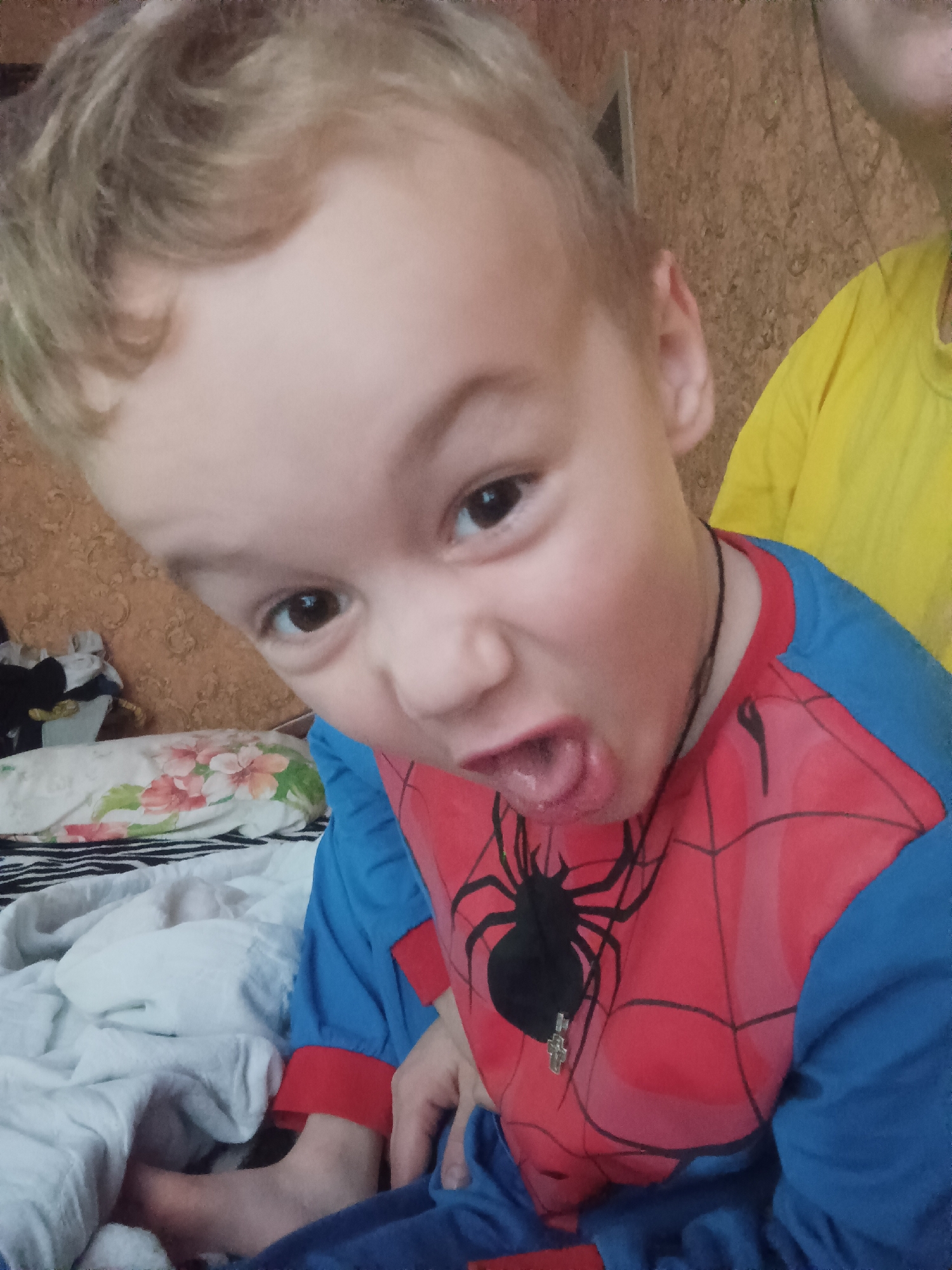 A child with light brown, wavy hair is wearing a blue and red Spider-Man-themed pajama top with a black spider graphic on the chest and a silver cross pendant on a black string around their neck. The child has brown eyes, pale skin, and is pulling a face, with their mouth open, showing their tongue. The child is sitting on a bed with a white, patterned pillow. In the background, there is a wall with a textured, brown pattern, and a person wearing a yellow shirt is partially visible. The location is likely an indoor space in Ilskiy, Russia, based on the available information.