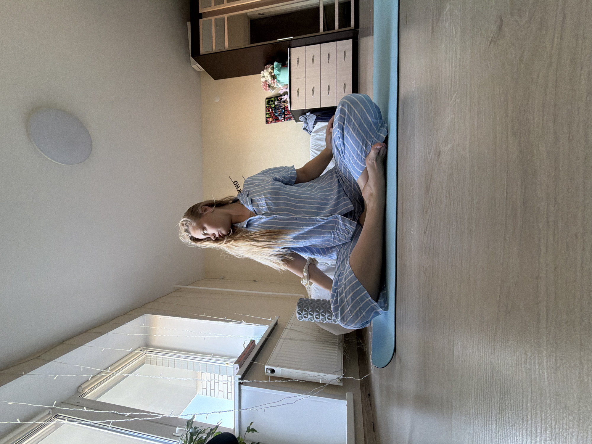 A woman is sitting in a cross-legged position on a blue yoga mat in a room. She is wearing blue and white striped pajamas. The room appears to be an apartment or a home in Izhevsk, Russia, based on the style of furniture and decor. There is a window with strings of lights hanging around it. A radiator is visible near the window. In the background, there's a closet or storage unit, some decorative items on a wall, and part of a bed. The floor is wood-look laminate. A circular light fixture is present on the ceiling.