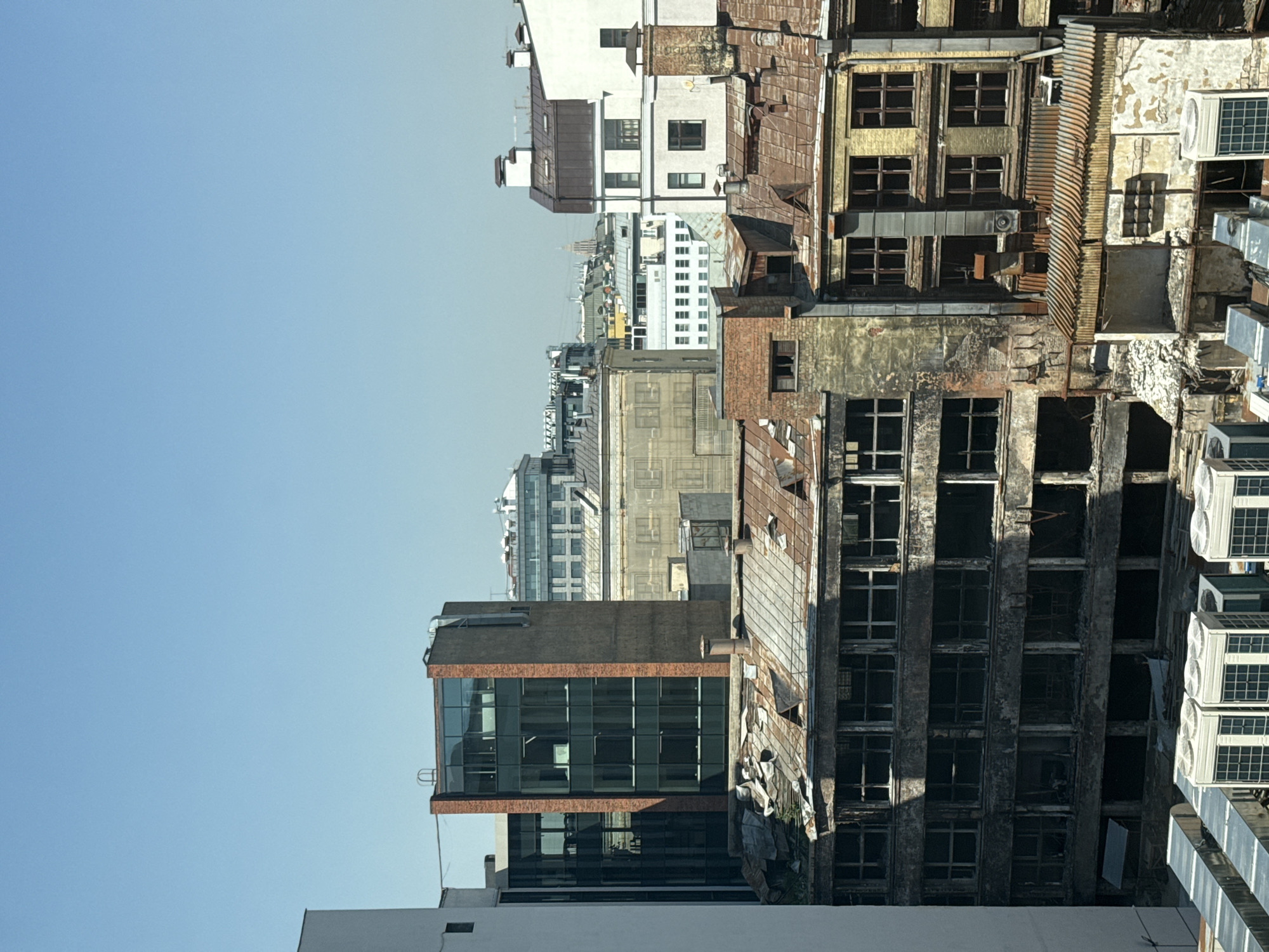 The image depicts a cityscape in Moscow, Russia, showcasing a mix of architectural styles and building conditions. In the foreground, a modern glass-walled building stands adjacent to an older, dilapidated structure with missing windows and decaying facade. The image captures the contrast between renovated and neglected urban spaces. Air conditioning units are visible on the exterior of the older building. In the background, more buildings, including white structures, stretch towards the horizon under a clear, blue sky. The scene is presented in a slightly tilted, vertical perspective.