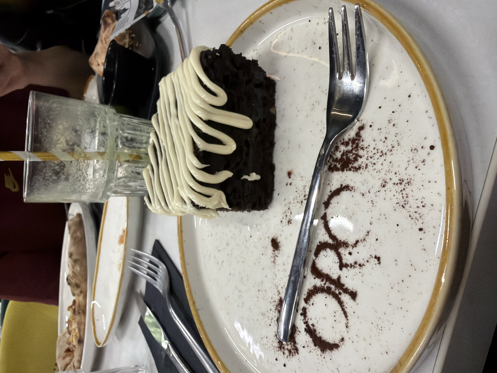 The image shows a plate of chocolate cake with white frosting, served with a fork, in what appears to be a restaurant. The cake is on a speckled plate with a gold rim. Beside the fork is a cocoa powder inscription "ORO". There are other plates with food and glassware visible, suggesting the setting of a meal. The location is potentially Panagyurishte, Bulgaria.