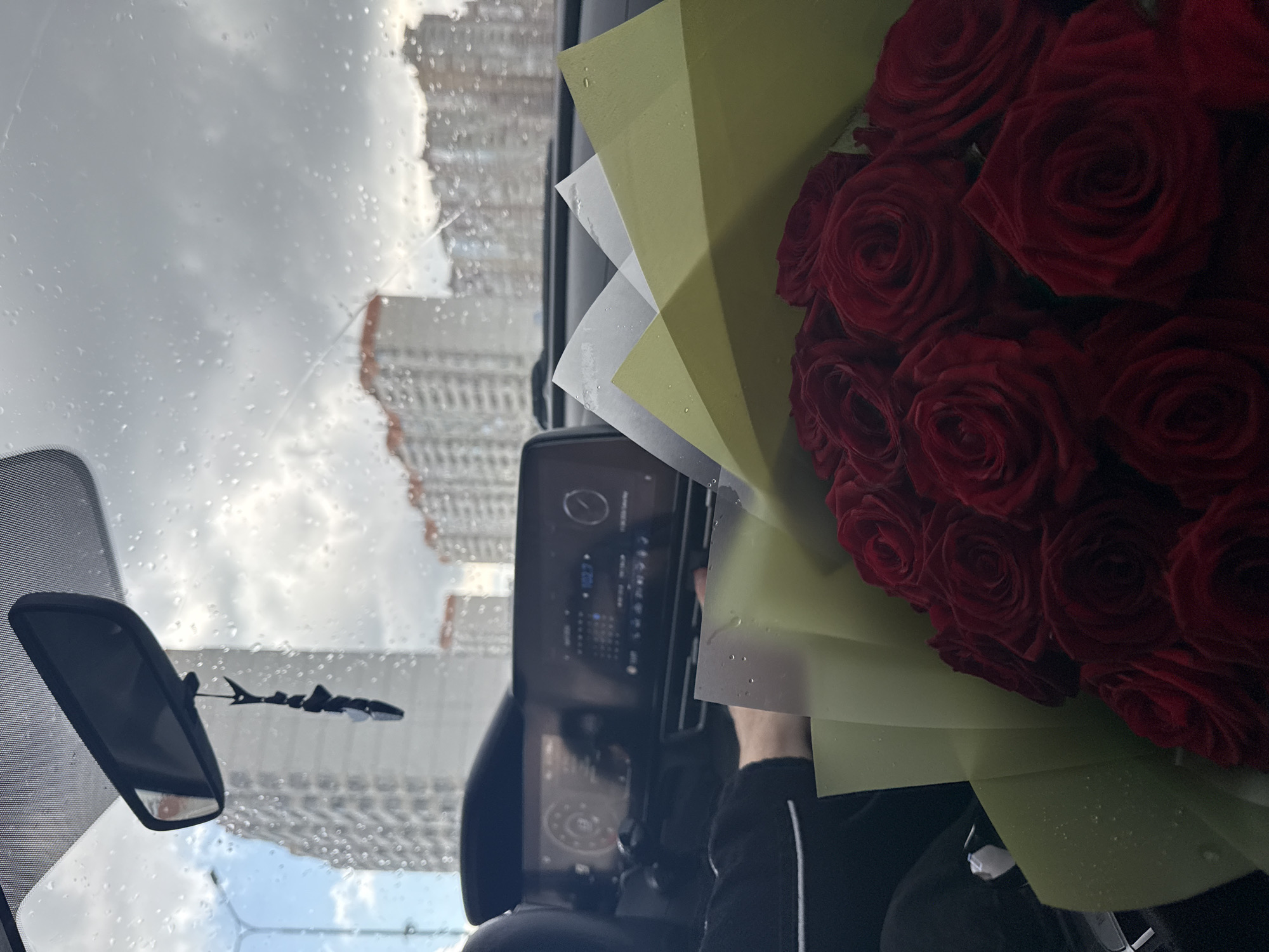 The image captures a view from inside a vehicle, possibly a car, looking out at a rainy cityscape. The windshield is covered in raindrops, and a rearview mirror is visible on the left. Through the windshield, tall buildings can be seen, indicating a densely populated urban environment. A bouquet of red roses is positioned on the right side of the frame, wrapped in green and white paper. A person's arm and part of their body can be seen holding the bouquet. The vehicle's dashboard and infotainment screen are also visible. The location is an unknown city, unknown country.