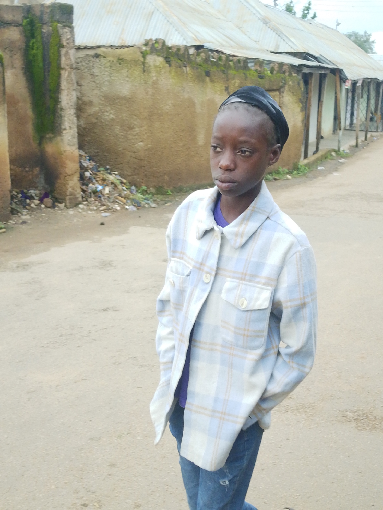 A young person wearing a black head covering and a light blue and white plaid button-down shirt over a purple t-shirt stands on a dirt road. Their hands are in their pockets. Behind the person is a building with a corrugated metal roof and walls made of mud or concrete, partially covered in moss and discolored with age. A pile of trash is visible near the base of the building. The scene appears to be in an urban or semi-urban environment, possibly a residential area, in Jos, Nigeria.