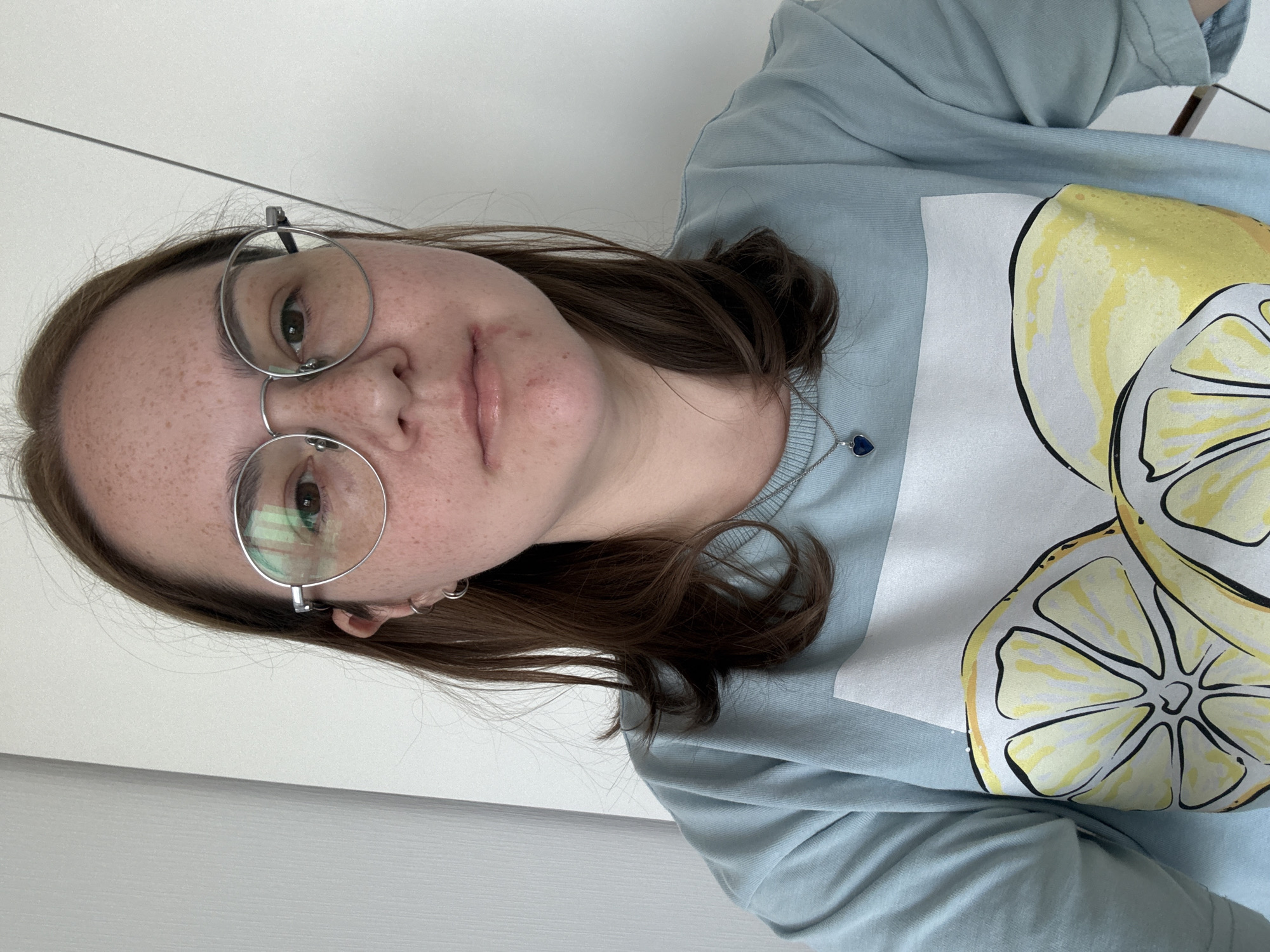 A medium shot photograph shows a young woman with light brown hair, wearing round, silver-rimmed glasses and a light blue t-shirt with a graphic print of sliced lemons. She has fair skin with freckles and a small heart-shaped pendant necklace. The background appears to be an indoor setting with a white wall, possibly in Lyubertsy, Russia.