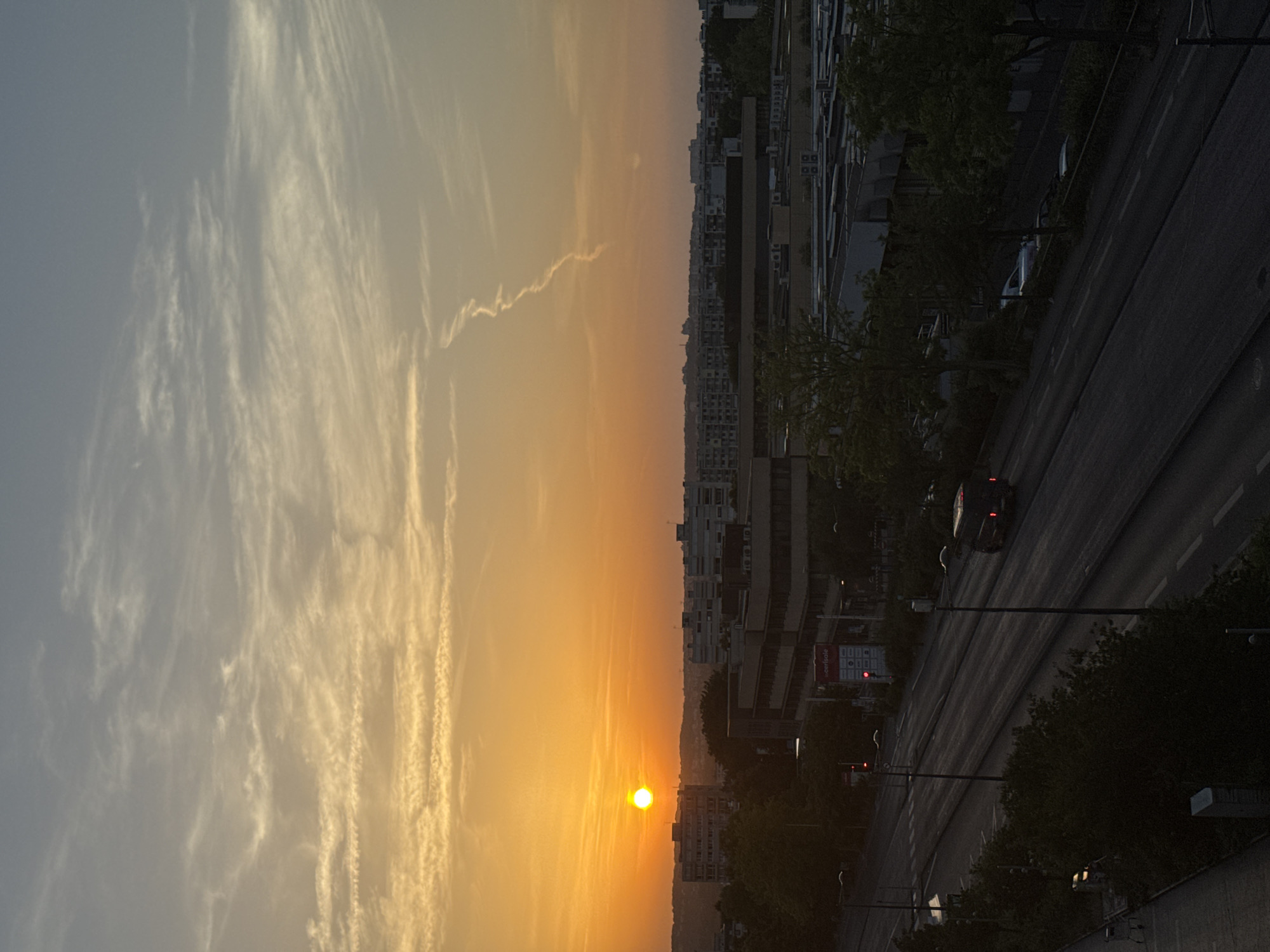 A vertical, high-angle shot captures a city skyline at sunset. The sun, a bright orange orb, is positioned in the upper left quadrant of the image, casting a warm glow across the sky and illuminating wispy, elongated clouds. The sky transitions from a pale orange near the horizon to a soft blue towards the top. Below the sky, a dense urban landscape unfolds, characterized by a series of buildings of varying heights and architectural styles, some appearing to be residential and others commercial or industrial. Trees are interspersed throughout the scene, their dark foliage contrasting with the brighter sky. A street with multiple lanes runs through the lower portion of the image, with a dark-colored van visible driving on the right side. Traffic lights are present on the street, with one red light illuminated. The overall impression is one of an urban environment during a transitional period of the day, with the natural beauty of the sunset juxtaposed against the man-made structures of the city.