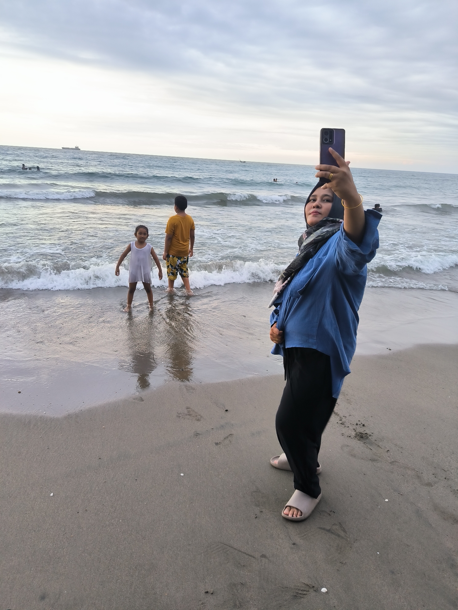 This detailed image depicts a beach scene in Lhokseumawe, Indonesia, under an overcast sky. In the foreground, positioned on the right, an adult woman with dark hair covered by a black hijab and a patterned scarf takes a selfie with a dark-cased smartphone, held aloft in her extended right hand. She wears a loose-fitting blue long-sleeved tunic, black trousers, and light grey open-toed slides. A gold-colored ring is visible on her right ring finger, and a gold-colored bangle is on her right wrist. Her left hand is placed over her abdomen.
In the midground, two children are wading in the shallow ocean water. To the left, a younger child wearing a white sleeveless dress stands facing the viewer, with water at ankle depth. To the right, an older child, wearing a yellow t-shirt and patterned shorts, stands facing away towards the ocean, with water reaching their mid-calves.
The ocean displays multiple breaking waves near the shore, and further out, two ships are visible on the horizon line. The beach itself is composed of wet, light-brown sand, showing visible footprints and scattered small shells. The sky is uniformly light grey, indicating an overcast condition.