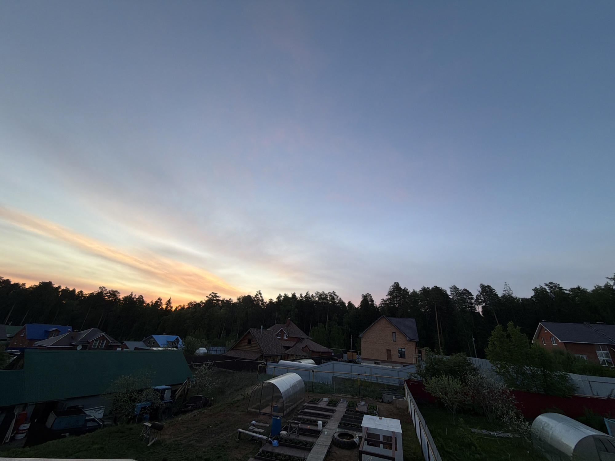 The image depicts a residential area near Chaikovsky, Russia, at dusk. The sky transitions from blue at the top to a gradient of light orange and yellow near the horizon, indicating the setting sun. A dark treeline is visible along the horizon, with multiple houses interspersed among the trees. In the foreground, there are various houses with different colored roofs (green, blue, and brown) and a plot of land with garden beds, greenhouses, and a white fence. The scene suggests a quiet, rural or suburban neighborhood. No people are visible in the image.
