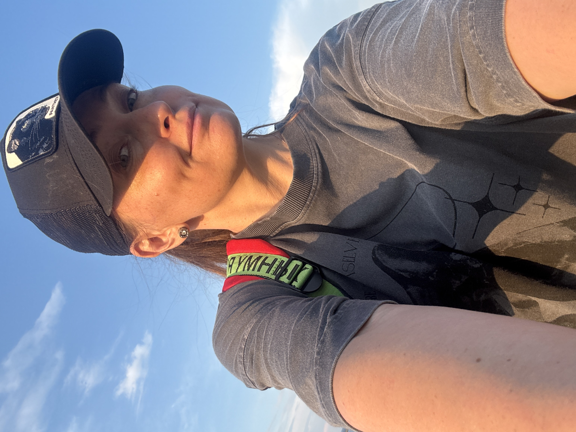 A woman is taking a selfie outdoors, with a bright blue sky and some clouds in the background. She is wearing a black baseball cap with a patch of a black panther on it, a gray t-shirt, and a backpack with a red and green strap. She has a small earring in her left ear. The lighting suggests it is either sunrise or sunset. The specific location in Chaikovsky, Russia, is not discernible from the image alone.