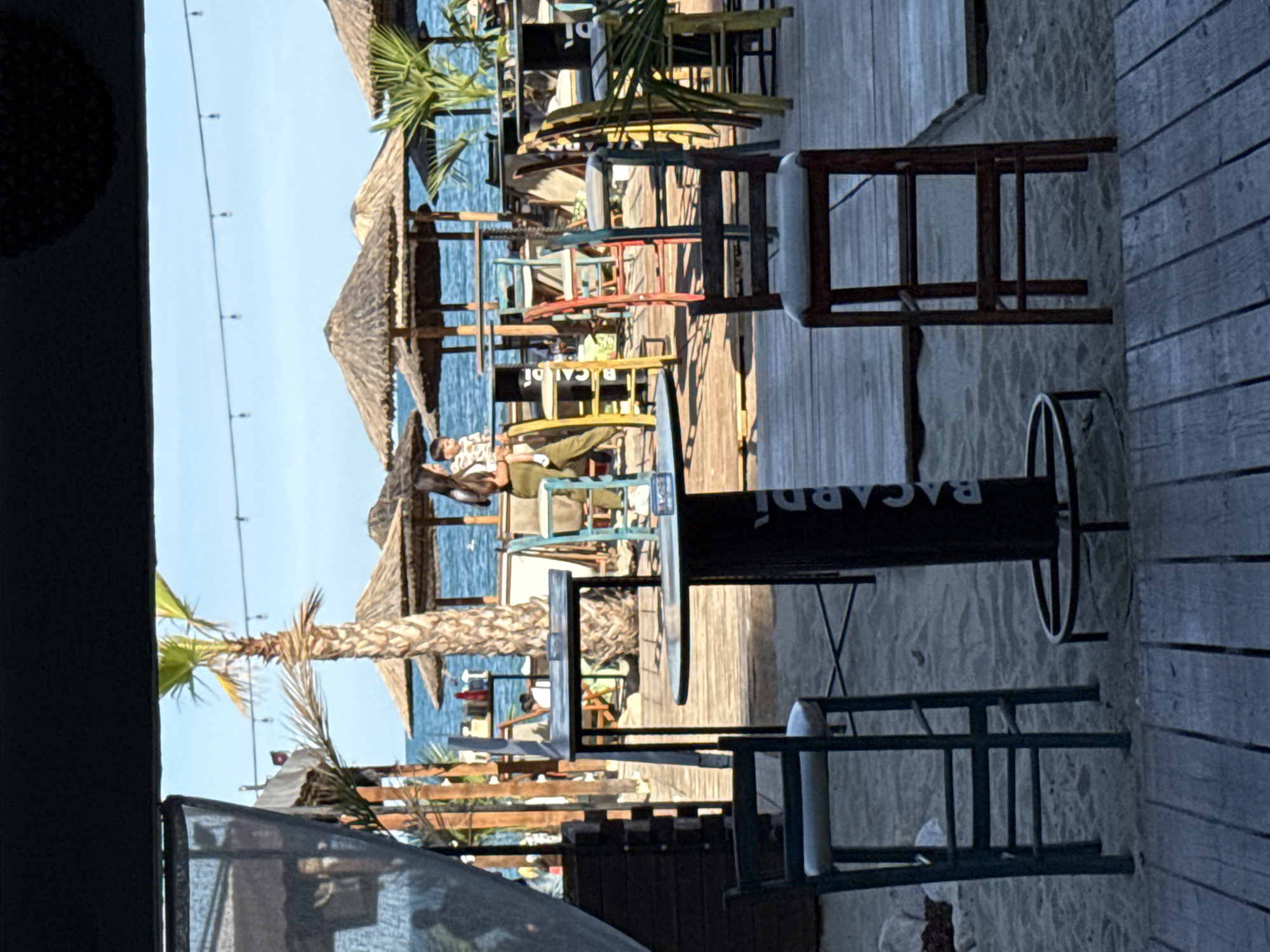 The image depicts a beachside bar or restaurant scene. In the foreground, there are wooden planks and sand, with several high stools and tables. A black table with the word "BACARDI" printed on it stands prominently. In the mid-ground, more colorful stools and tables are visible, leading towards the beach. Behind the seating area, there are thatched umbrellas providing shade on the sandy beach, and beyond that, the blue sea and sky are visible. A palm tree is located behind the Bacardi table. A few people are scattered in the background, near the umbrellas and the water, some appearing to be walking or standing. The overall ambiance suggests a relaxed, outdoor dining or drinking establishment by the sea.