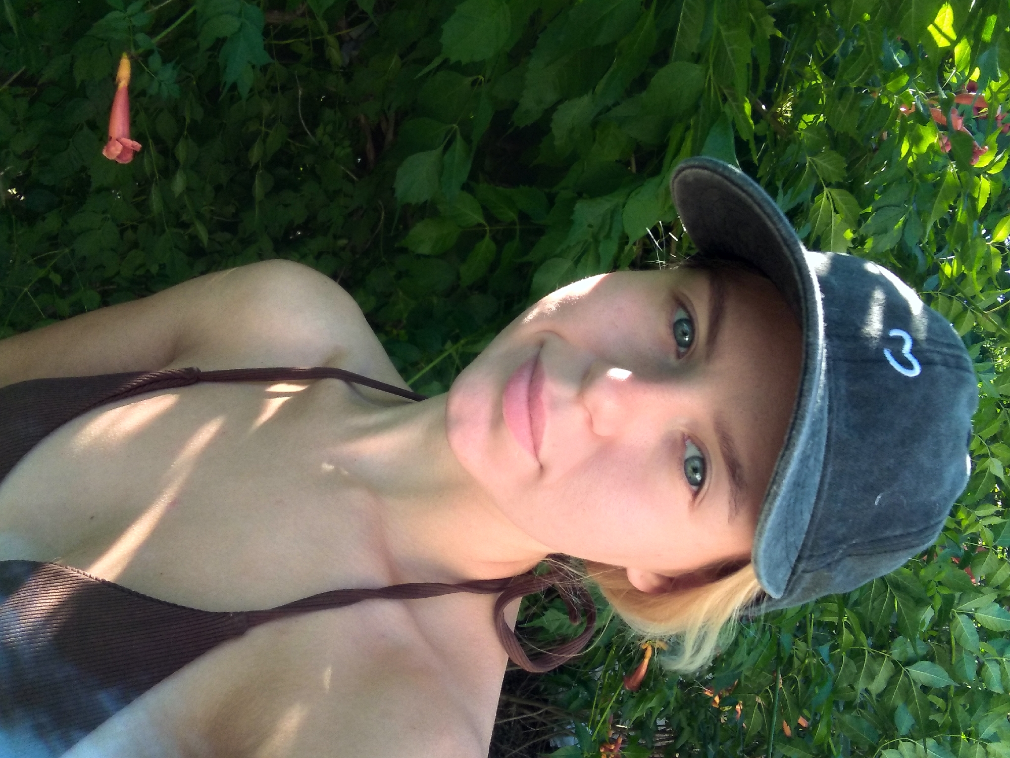A young woman with blonde hair, wearing a dark baseball cap with a white heart logo and a brown bikini top, is pictured outdoors. She is looking towards the camera, with greenery and foliage forming the background. A single, elongated pink flower is visible on the left side of the frame, among the leaves. The lighting suggests it is daytime, with sunlight casting shadows on her skin and clothes. The location is identified as Radisne, Ukraine.