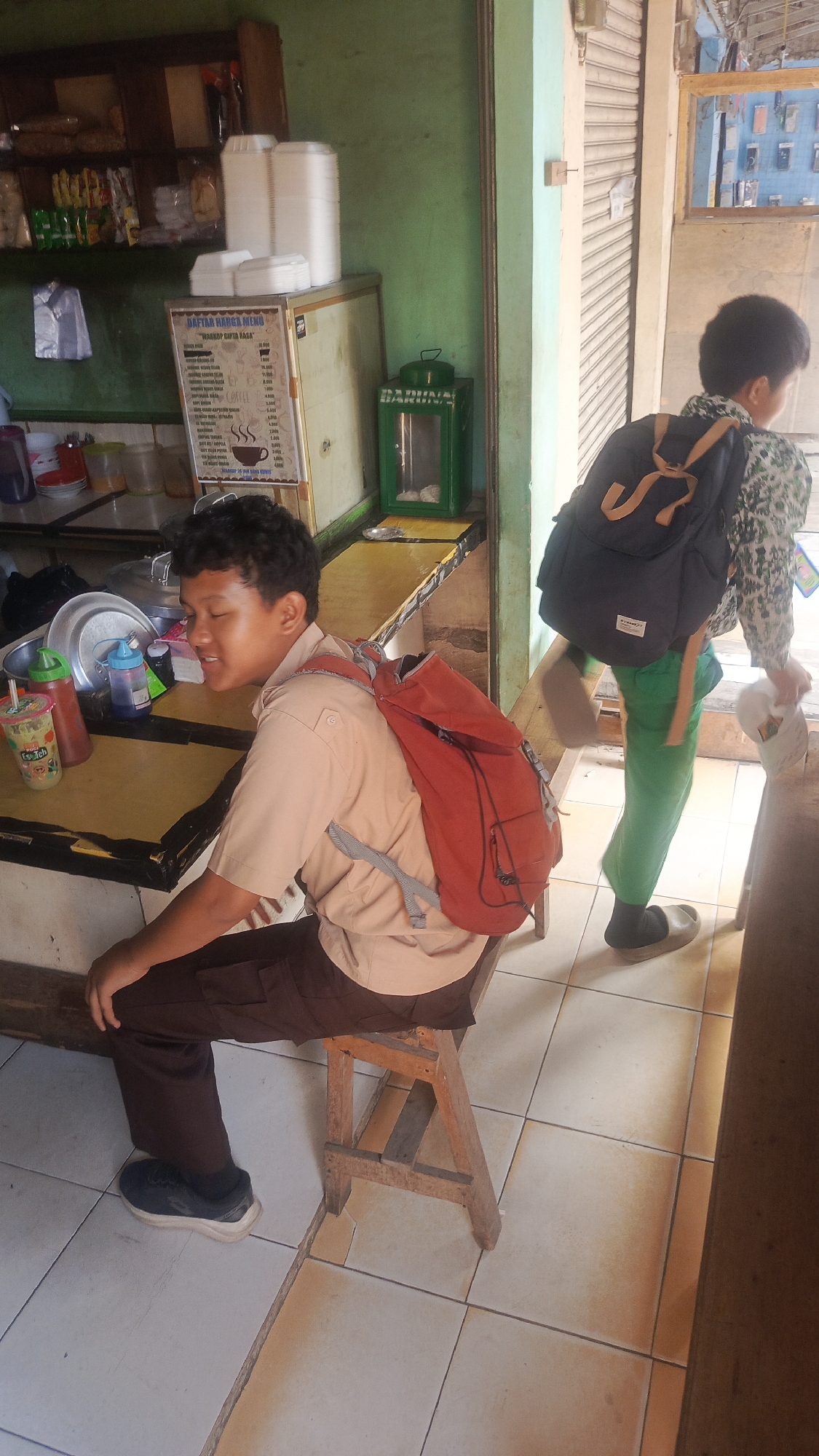 In Parung Panjang, Indonesia, two young boys, likely students, are seen in an indoor setting that appears to be a food stall or small shop. The boy in the foreground, wearing a light brown uniform shirt and dark brown pants, is seated on a wooden stool. He has a red backpack on his back and is looking to his right with his eyes closed. A tray of food items, including drinks in plastic cups and bottles, is visible on a yellow counter in front of him. To the right, a second boy, wearing a patterned shirt and green pants, walks away from the camera, carrying a dark blue backpack. The environment features a green wall, shelves stocked with packaged goods, and a metal display case. The floor is tiled with a light color. The scene captures a moment of daily life, possibly during a break or after school, in a typical Indonesian establishment.