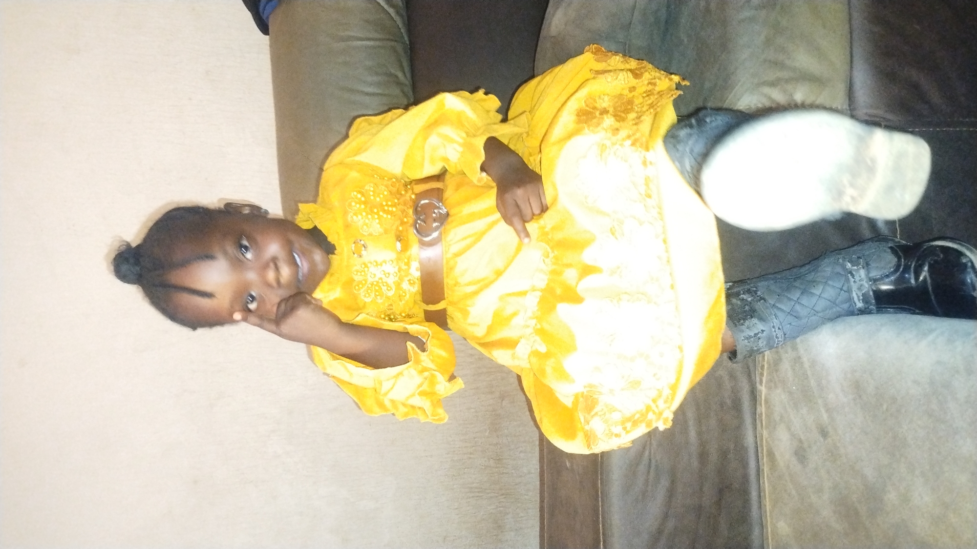 A young girl lies on a brown couch in what appears to be a residential setting. She wears a bright yellow dress with a brown belt. The dress features decorative embellishments on the bodice. She is wearing black boots. She makes an 'OK' gesture with her hand, looking at the camera. The exact location within Jos, Nigeria, is not discernible from the image.