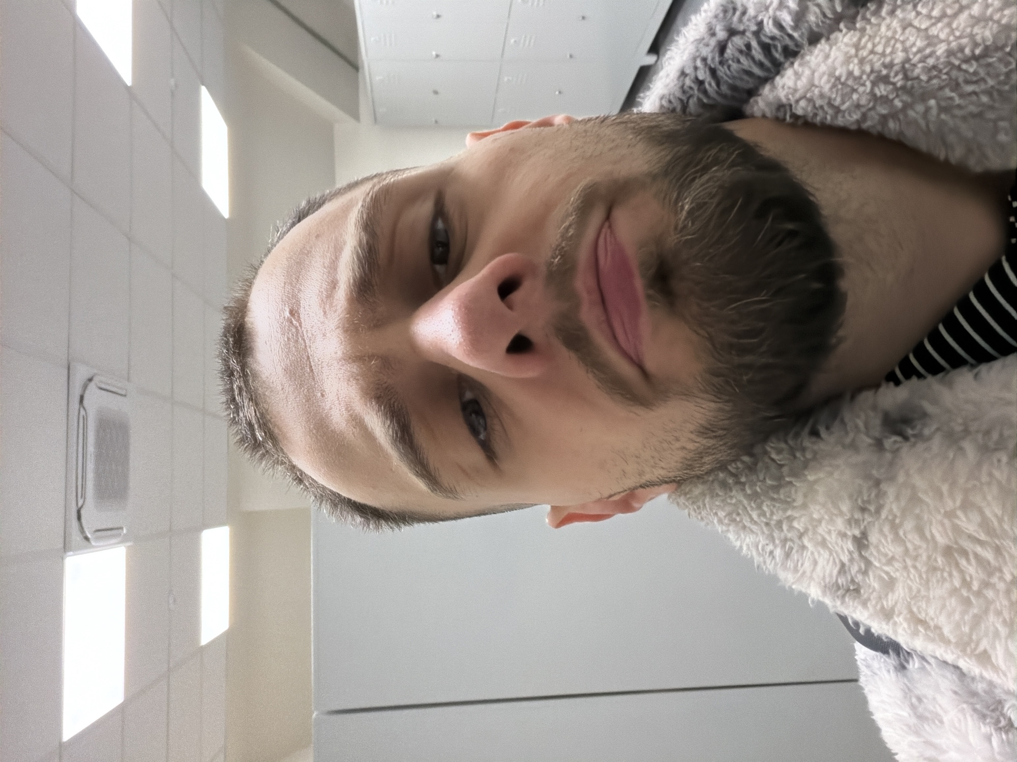A close-up, slightly tilted portrait of a man with short dark hair and a full beard. He has fair skin and is wearing a gray, fuzzy jacket with a black and white striped shirt underneath. The background appears to be a brightly lit, sterile environment, possibly a bathroom, with white tiled walls, rectangular lights, and a ventilation grate visible. The man's expression is neutral. No specific location in Plovdiv, Bulgaria is identifiable from the image.